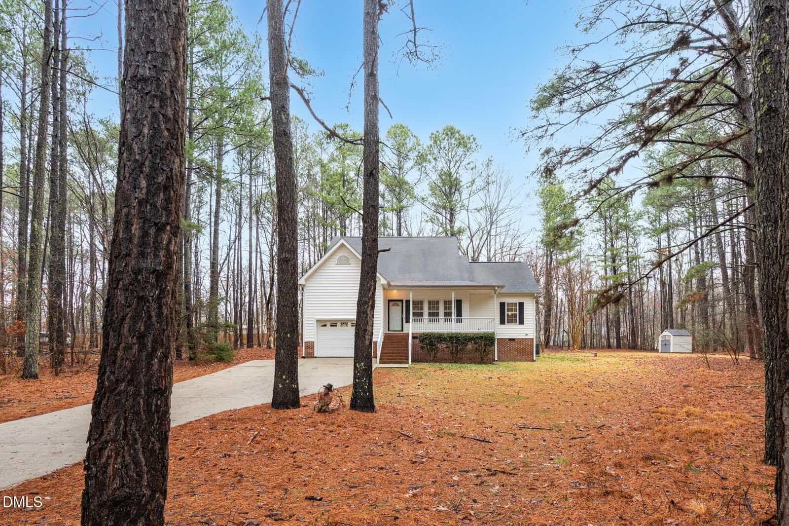 A white single-story house with a front porch and attached garage sits in a wooded area. A concrete driveway leads to the house. Tall pine trees surround the property, and a small shed is visible in the backyard.