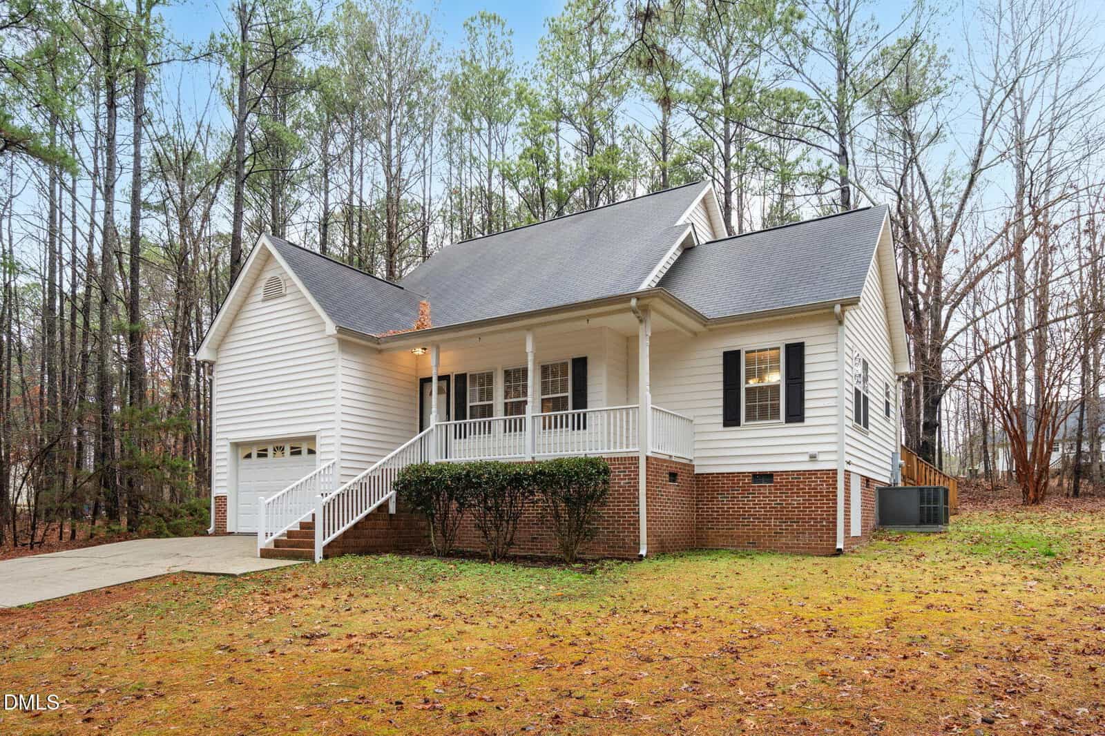 Single-story house with white siding, a brick foundation, front porch with white railings, attached garage, and a paved driveway, set among tall trees and surrounded by a natural, leaf-covered yard.