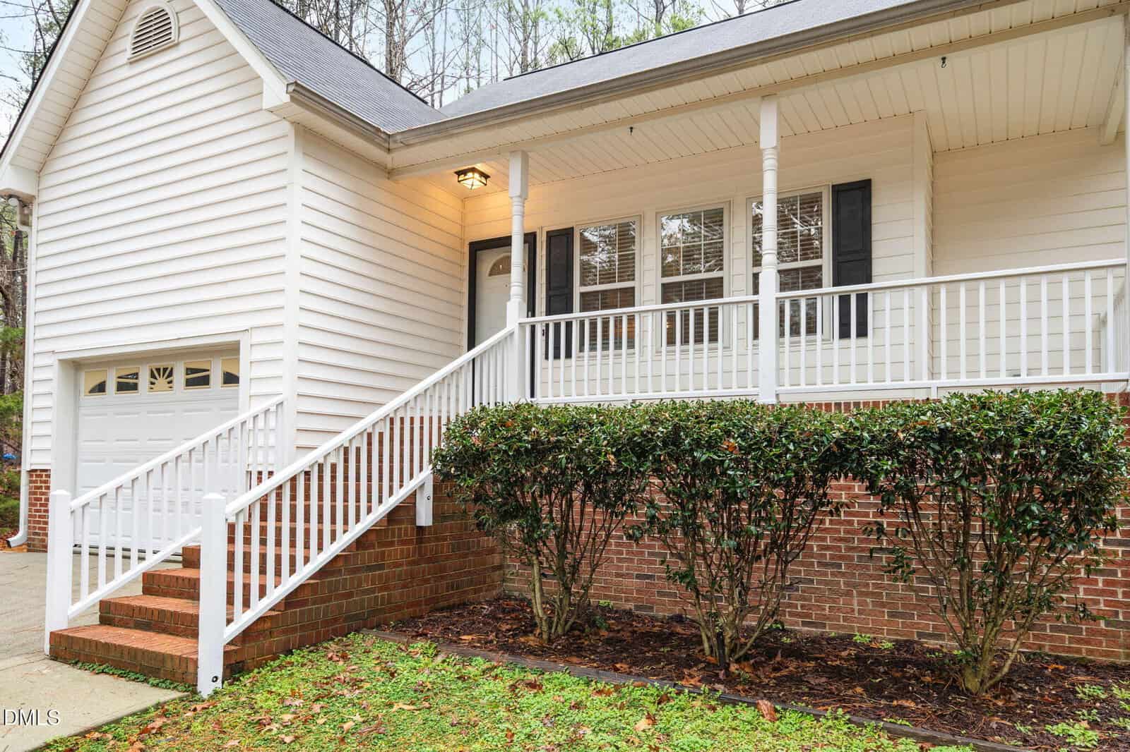 Single-story house with white siding, a covered front porch, white railing, brick foundation, attached garage, and neatly trimmed bushes in front. Concrete driveway and steps lead to the porch. Trees visible in the background.