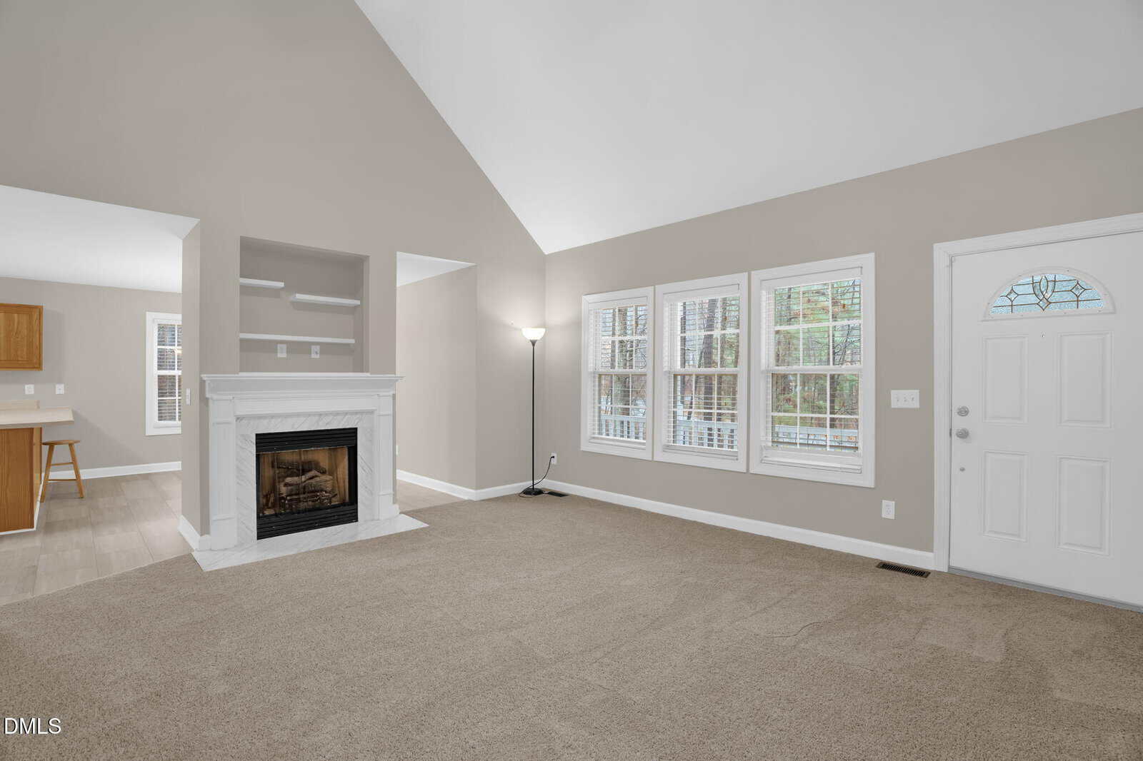 A spacious living room with beige carpet, light gray walls, a white fireplace, two large windows with blinds, a white door with a small arched window, and an adjacent open kitchen area.