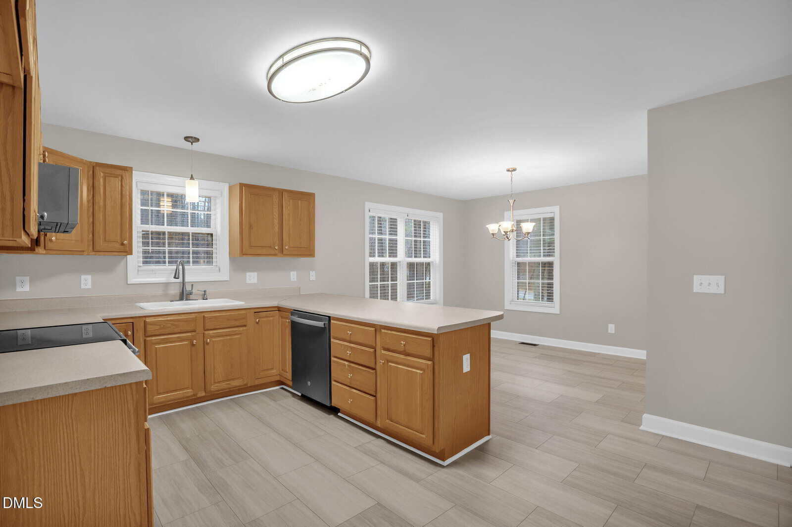 A spacious kitchen with light wood cabinets, light countertops, stainless steel appliances, and beige tile flooring. The room opens into a dining area with a chandelier and multiple windows.