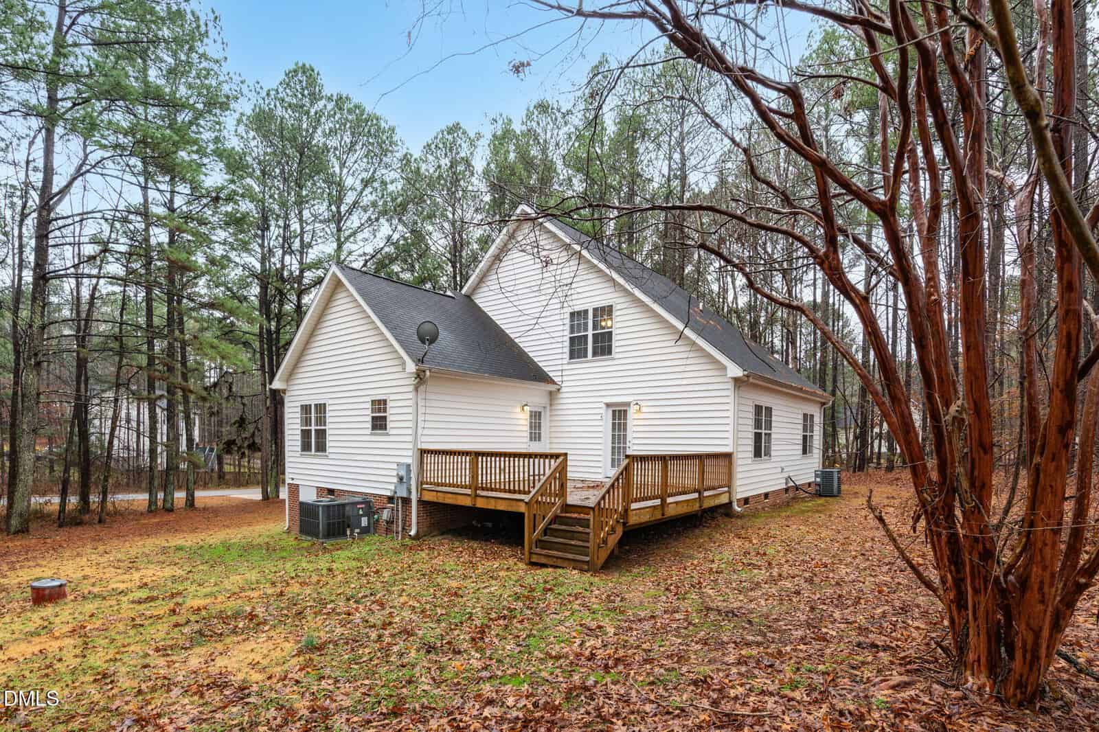 A white, two-story house with a wooden back deck is surrounded by trees and a yard covered in fallen leaves. The house has several windows, a satellite dish, and two outdoor AC units.