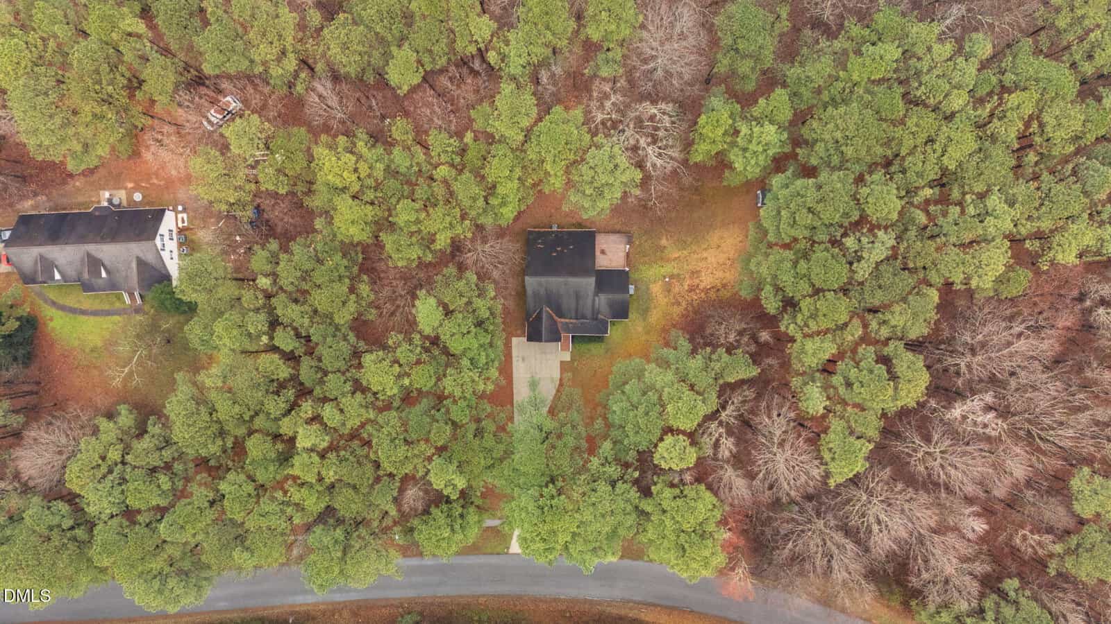 Aerial view of a house surrounded by dense trees, with a driveway leading to the road. Another house is partially visible on the left side. The area appears wooded and suburban.