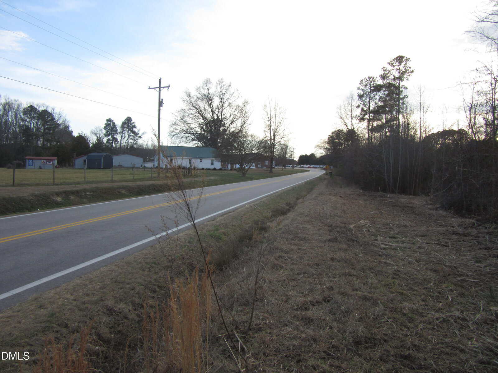 A two-lane road curves through a rural area, bordered by grassy shoulders and trees. Utility poles run alongside the road, and a few buildings are visible in the background under a partly cloudy sky.