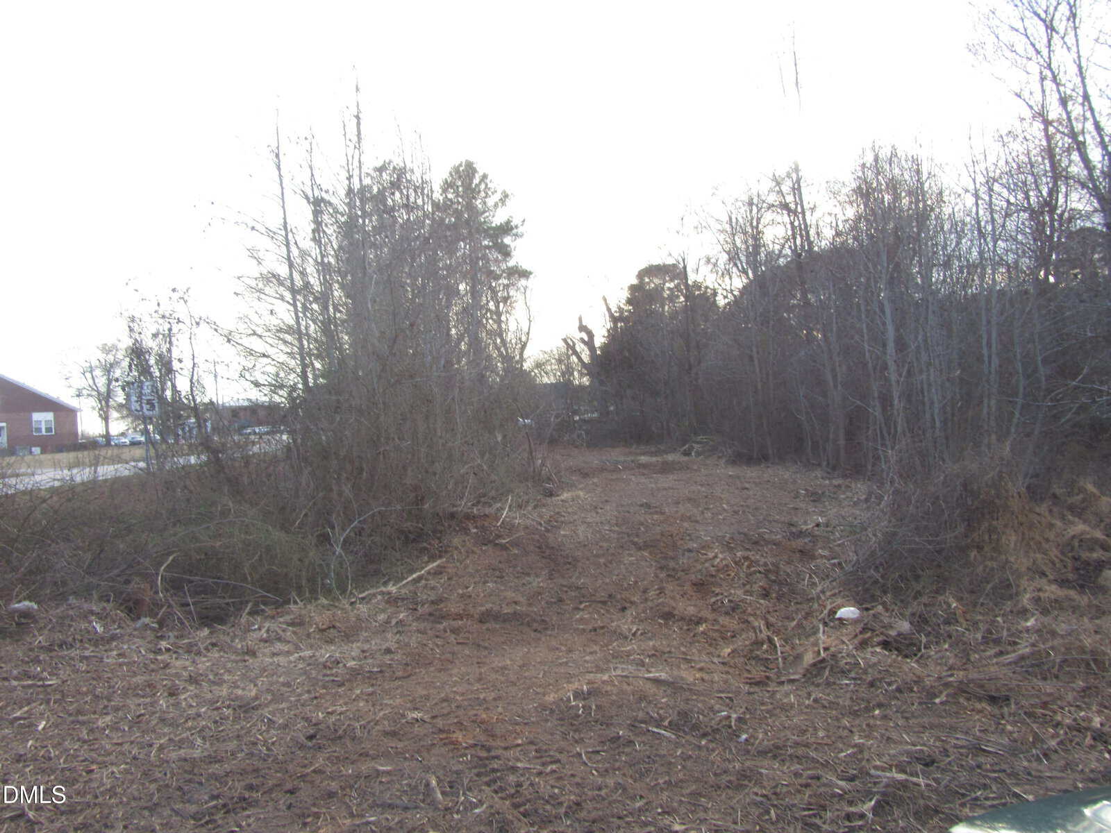 Cleared dirt path runs through a mostly bare, wooded area with leafless trees. Sparse brush and branches line both sides. A building and a road are visible on the left side in the background.