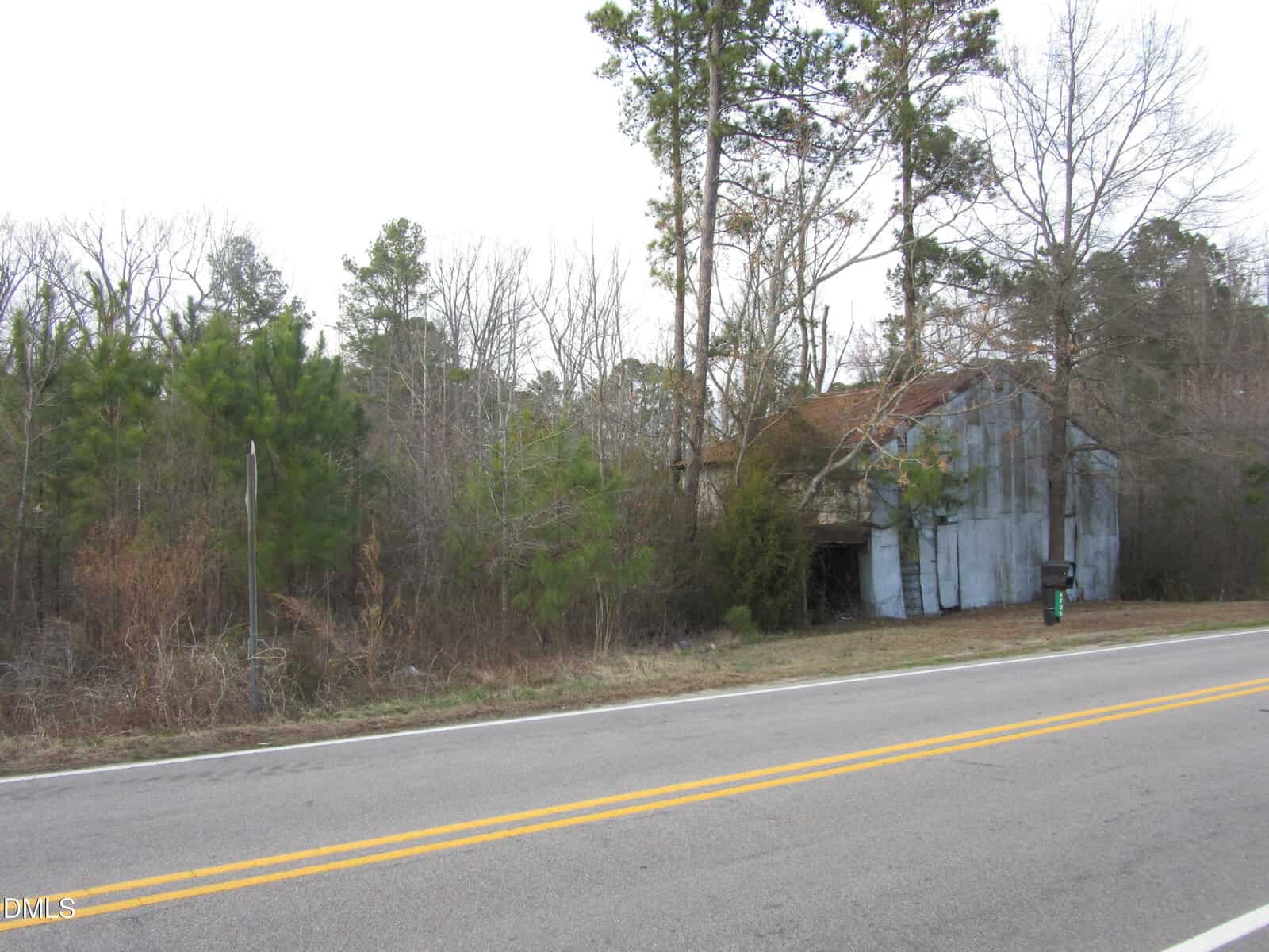 An old, weathered metal barn stands beside a rural two-lane road. The structure is partially obscured by overgrown trees and bushes, and the sky is overcast. A green mailbox is visible near the barn.