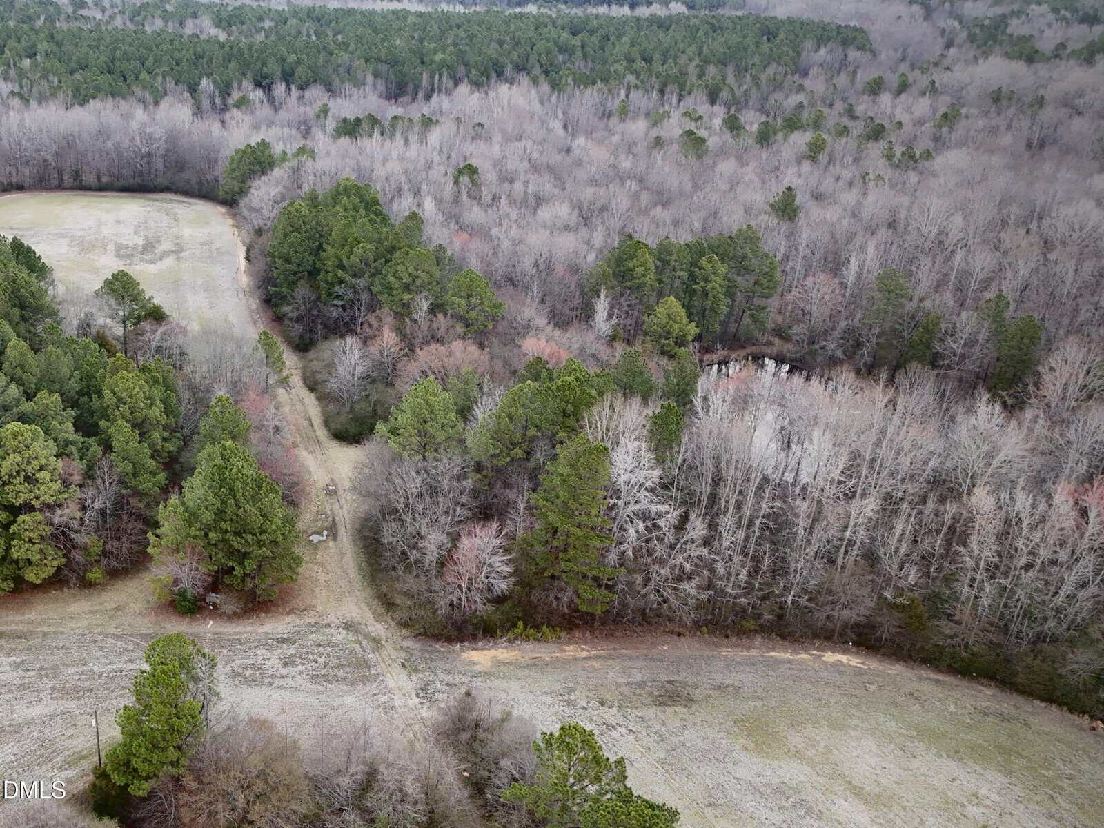 Aerial view of a rural landscape with a dirt path winding through open grassy fields and dense, leafless and evergreen trees, on an overcast day. A small pond is partially visible among the trees.