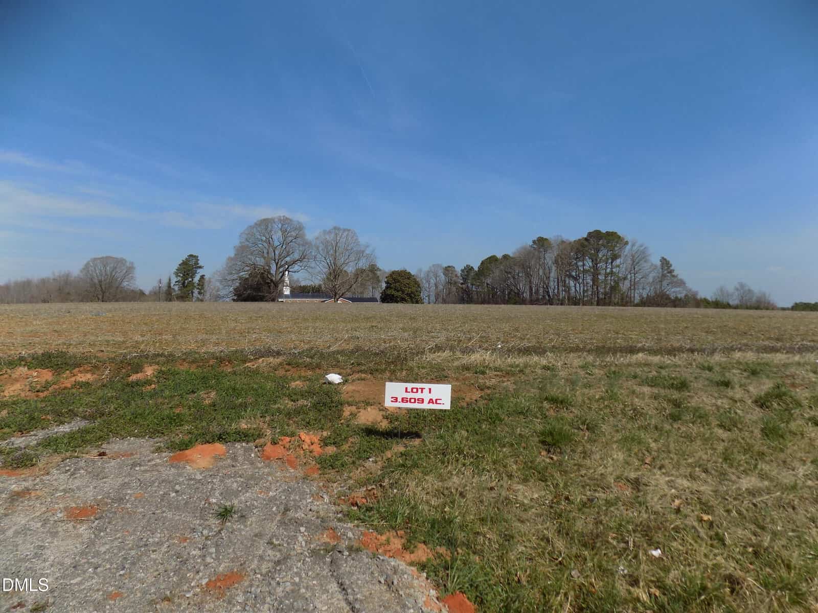 Open grassy lot on 0 Sutton Road in Louisburg, with a small red and white sign reading “LOT 1 3.609 AC.” in the foreground. Bare trees and a building are visible in the distance under a clear blue sky.