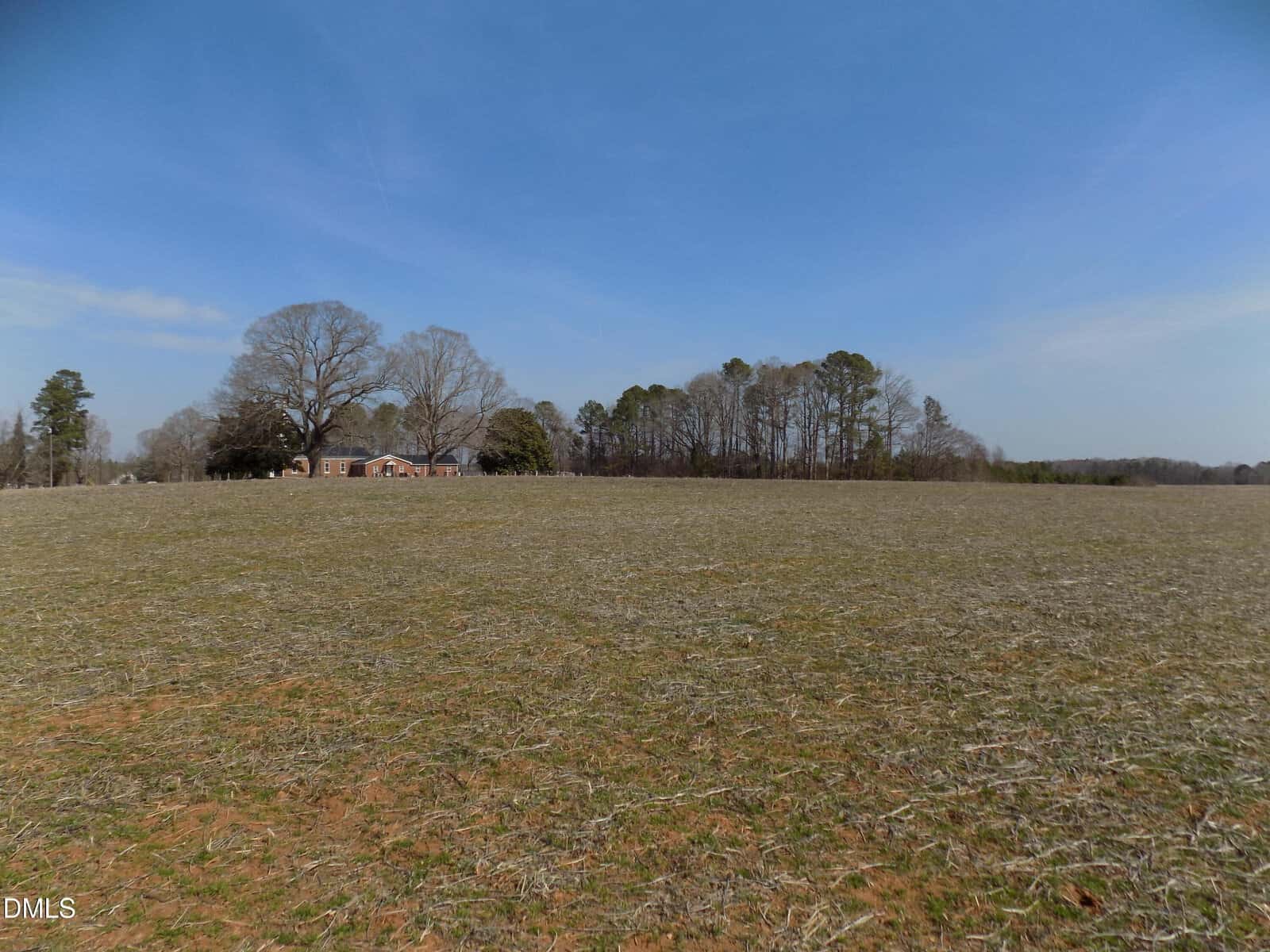 A large, open field with sparse grass and a red brick house surrounded by trees in the distance under a clear blue sky.