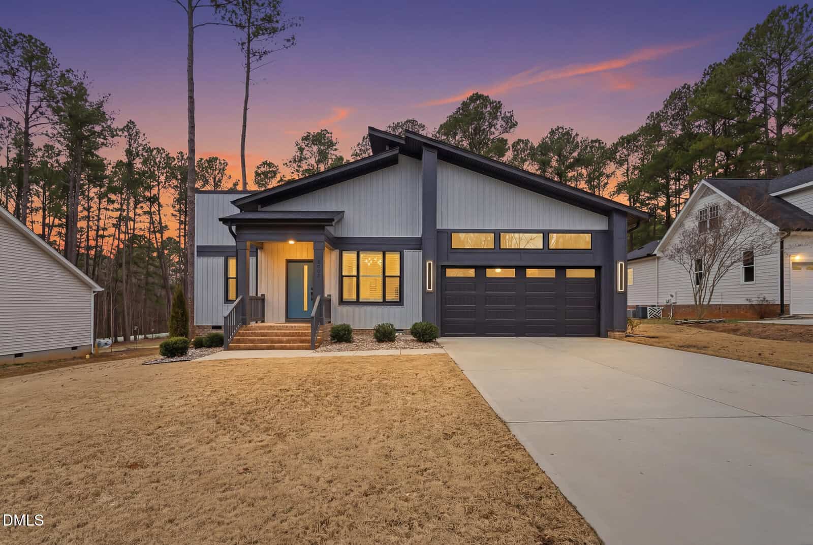 Modern single-story house with dark trim, large windows, and an attached two-car garage located at 2662 Sledge Road in Louisburg. The front yard has dry grass, a concrete driveway, and a small porch. Trees and neighboring houses are visible at sunset.