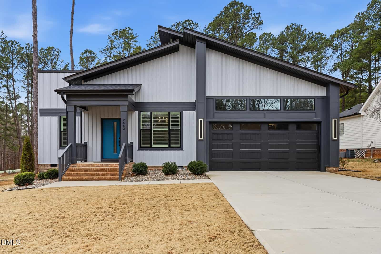 A modern single-story house with gray siding, black trim, a teal front door, large windows, a double garage, and a concrete driveway, set against a backdrop of trees and a dry, yellowish lawn.