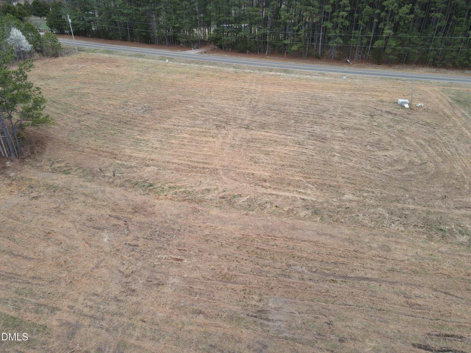 A large, cleared field with brown grass and parallel tire tracks sits beside 2 Coley Road in Henderson, bordered by a road and dense trees in the background under an overcast sky.
