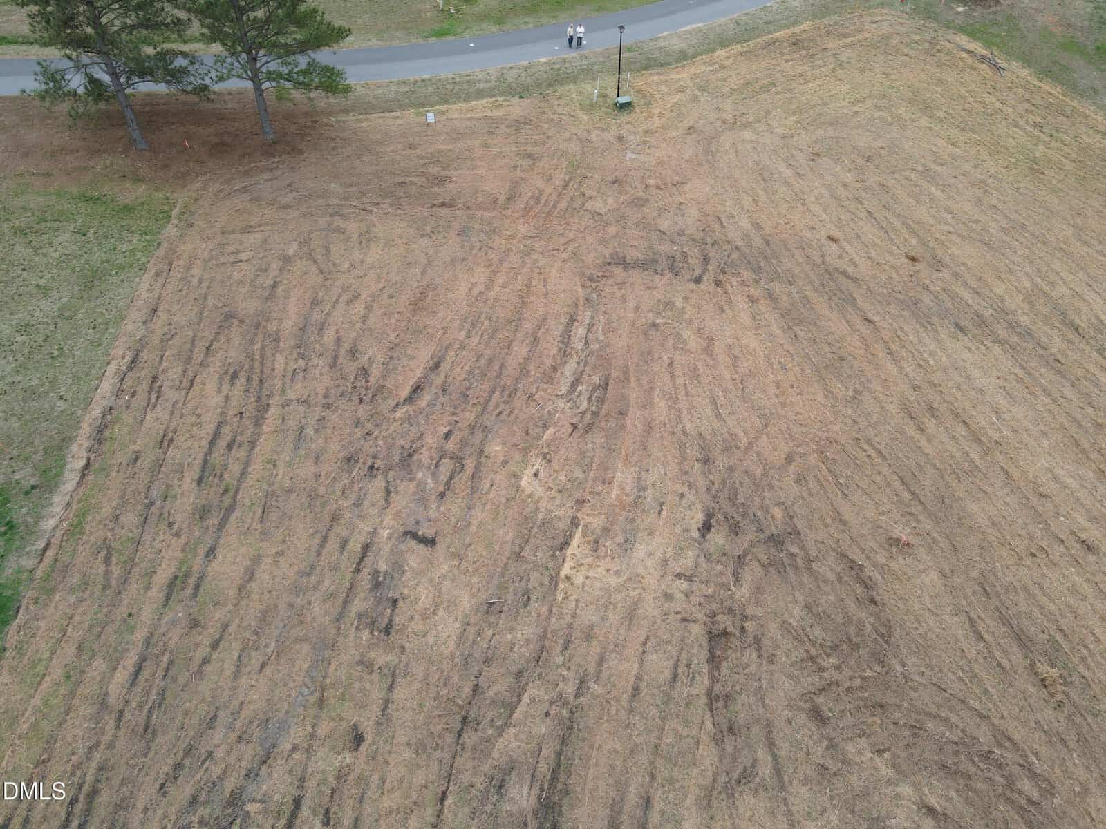 Aerial view of Lot 21 Sandy Plains Lane, Henderson—a large, cleared, brown grassy lot with visible tire marks. Two people stand near a road and some trees at the top edge of the image.