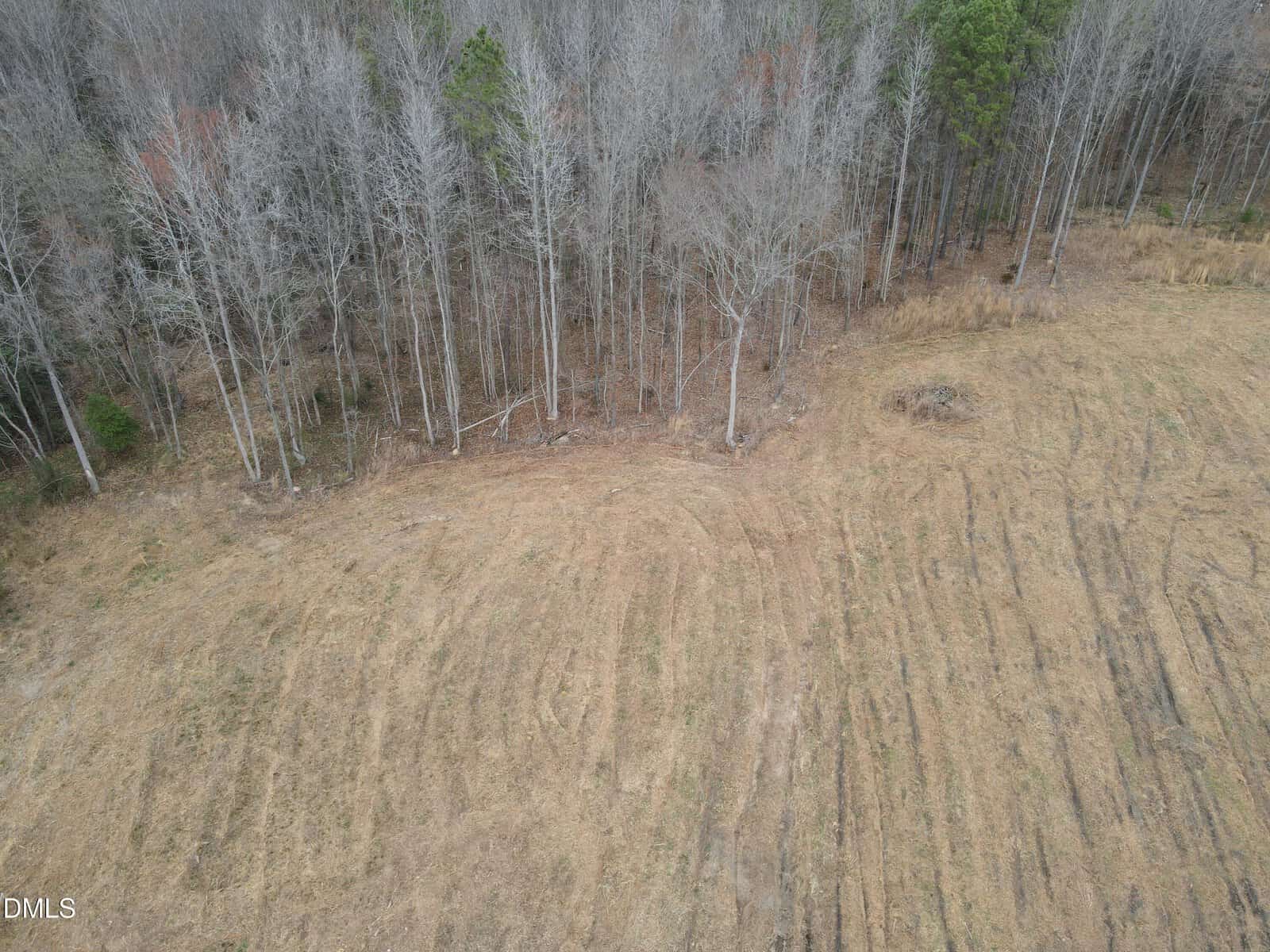 Aerial view of a barren field meeting a line of leafless trees at the edge of a forest, with patchy grass and visible tire tracks on the ground.
