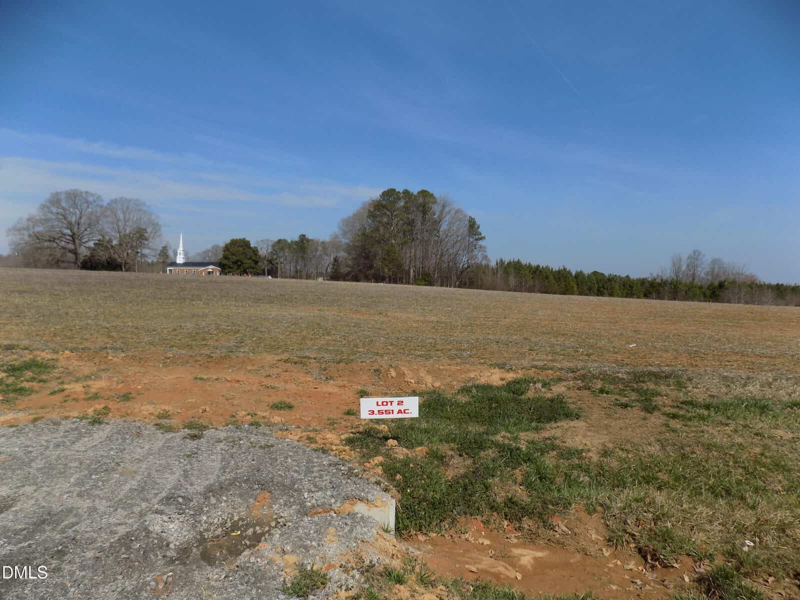 A mostly flat, empty lot with sparse grass and patches of exposed soil sits just off 00 Sutton Road in Louisburg. A small sign in the foreground reads “LOT C 3.551 AC.” Trees and a building with a white steeple are visible in the background.