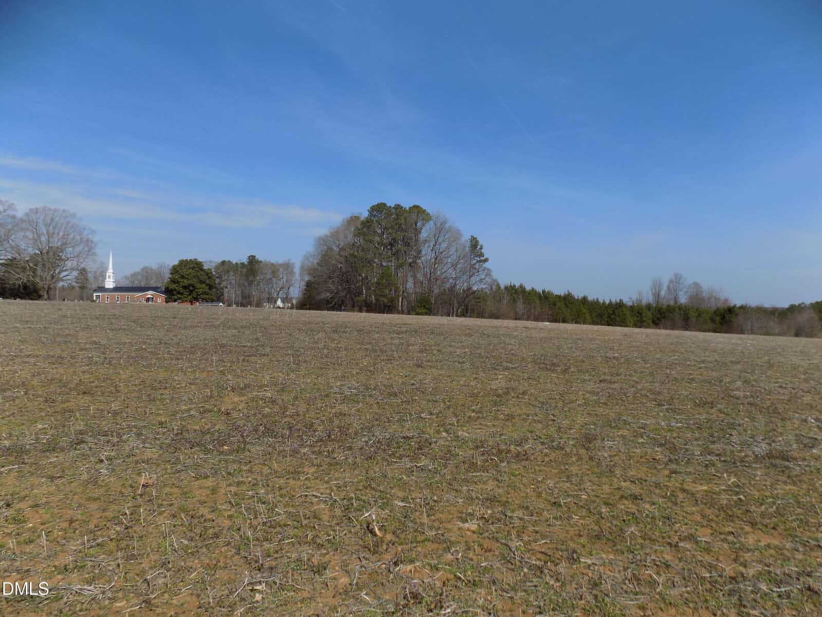 A large, mostly barren field stretches toward the horizon, bordered by leafless and evergreen trees. A white church with a tall steeple is visible in the distance under a clear blue sky.