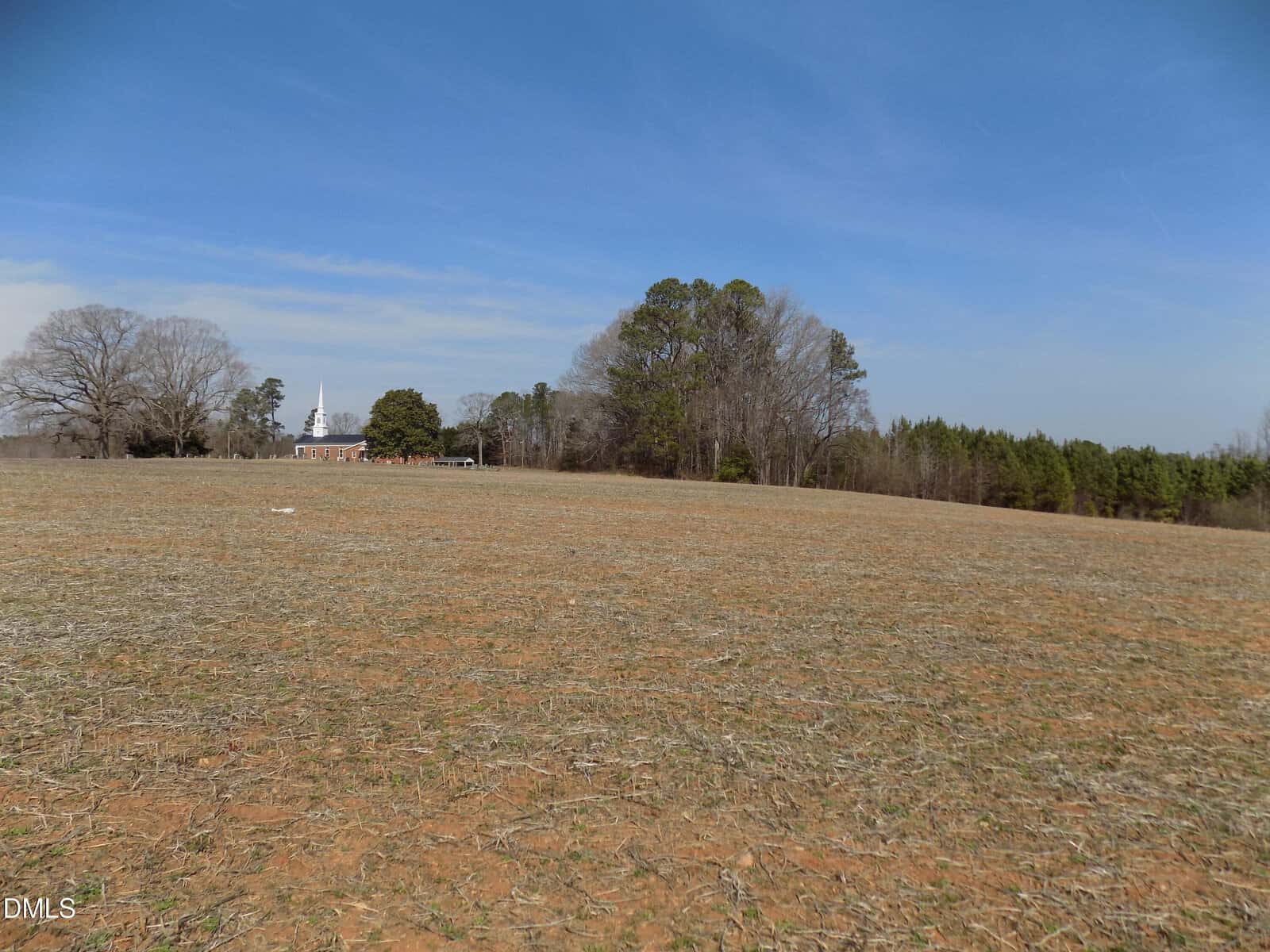 A wide, mostly barren field with sparse grass, a cluster of trees, and a distant building with a white steeple under a blue sky. The landscape is flat and rural.