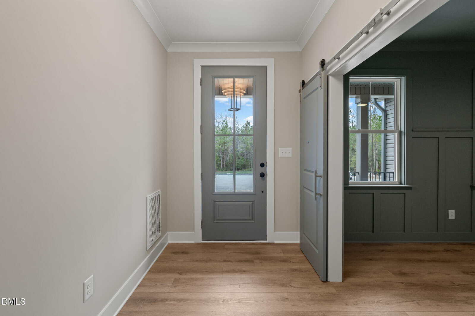Entryway with a glass-paneled gray front door, light wood flooring, and a sliding barn door opening to a dark-paneled room with a window; outside, trees are visible through the door and window.