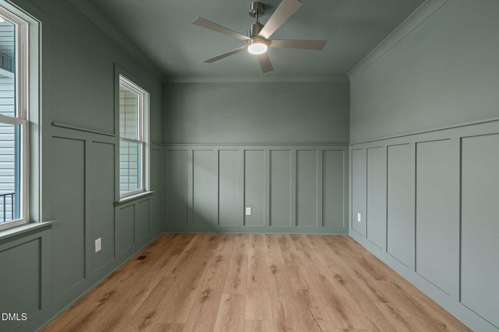 Room with green paneled walls and ceiling, light wood floor, two windows on the left, and a modern ceiling fan with light in the center. The space is empty and brightly lit by natural light.