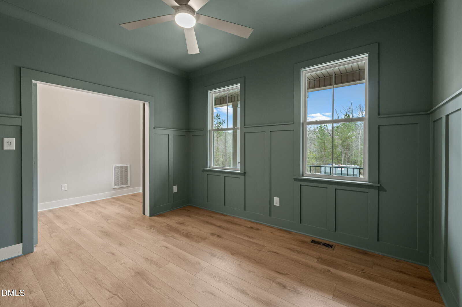 Spacious room with green wainscoted walls, light wood floors, a ceiling fan, and two windows showing an outdoor view. An open doorway leads to an adjacent, light-colored room.