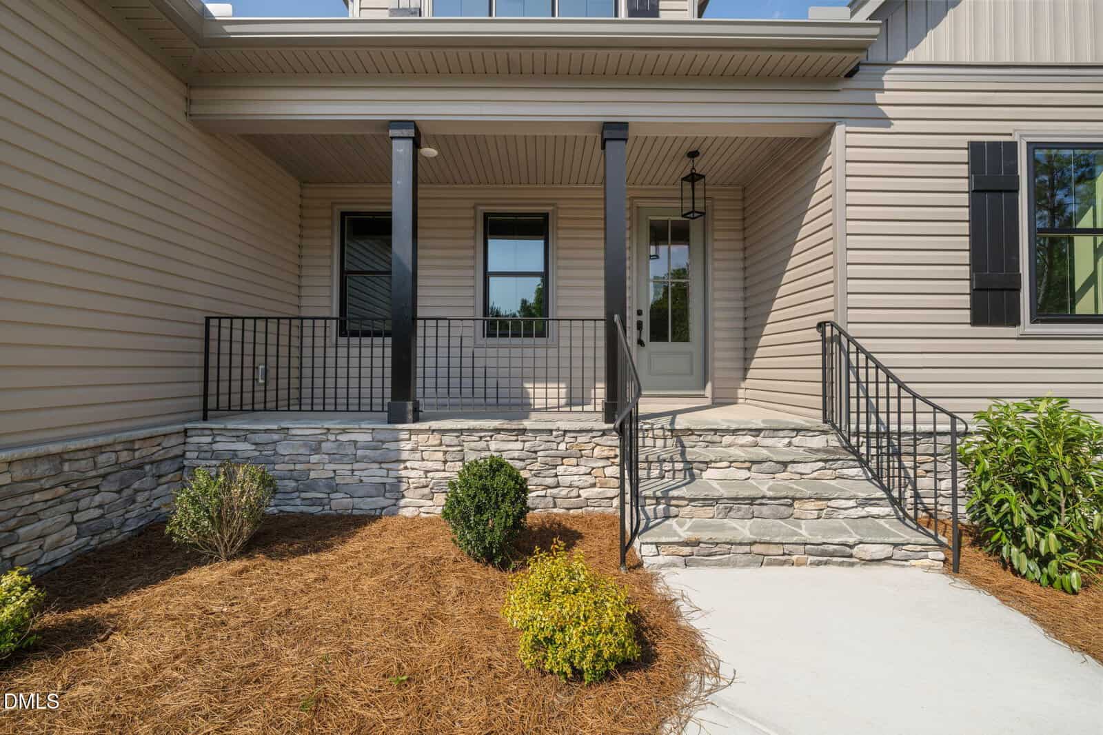 A stone walkway with steps leads to a covered porch with two black columns and a glass door. The exterior walls are beige with black window shutters. Small shrubs and mulch line the landscaped area near the entrance.