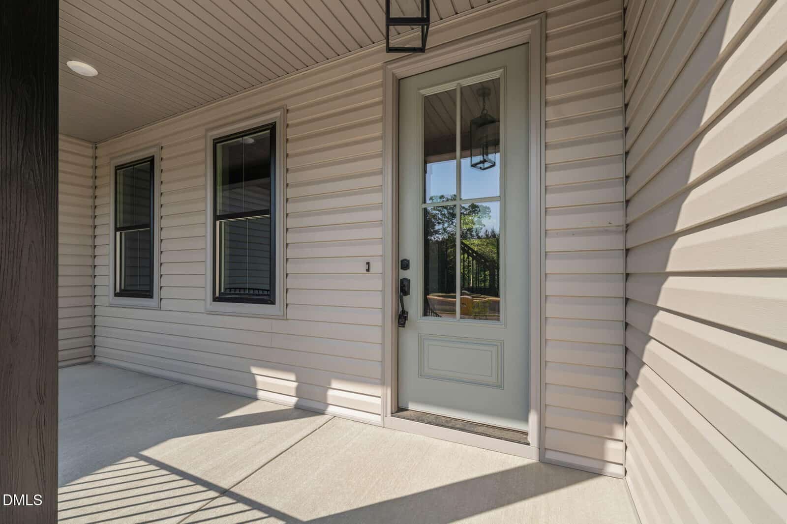 Covered front porch with beige siding, a glass-paneled door with black hardware, two black-trimmed windows, and a hanging lantern light fixture above the entrance. Sunlight casts shadows on the concrete floor.