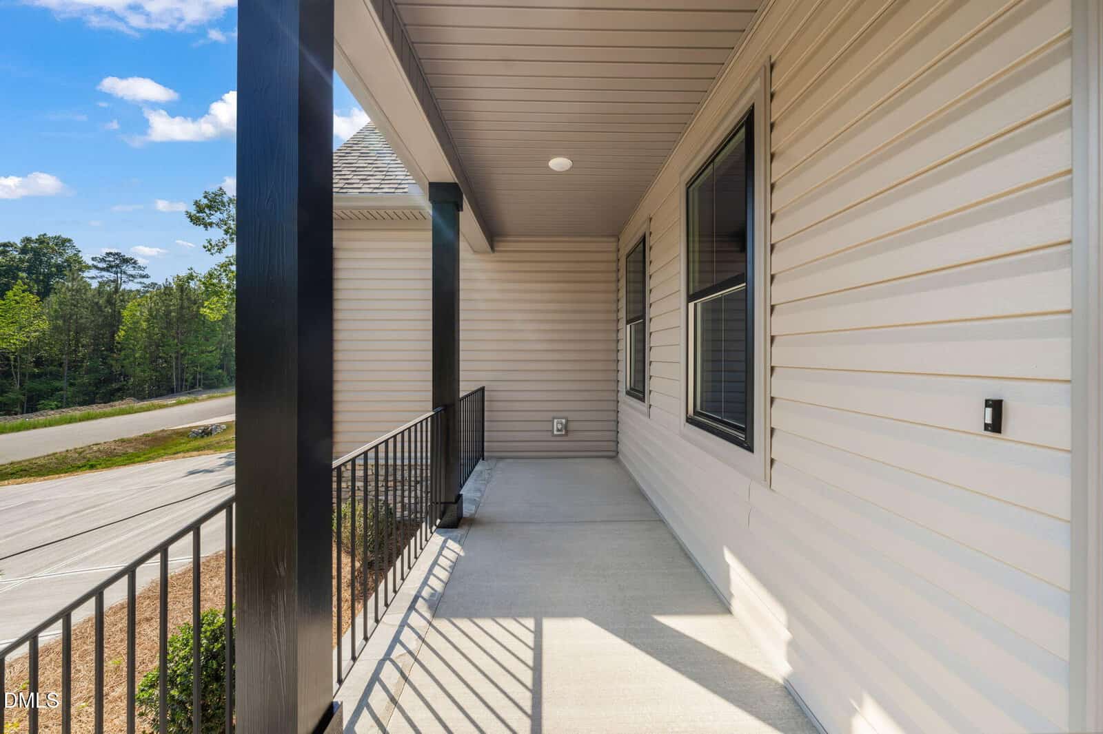 A covered front porch with beige siding, black metal railings, and a concrete floor. The porch overlooks a quiet street and green trees under a blue sky with scattered clouds.
