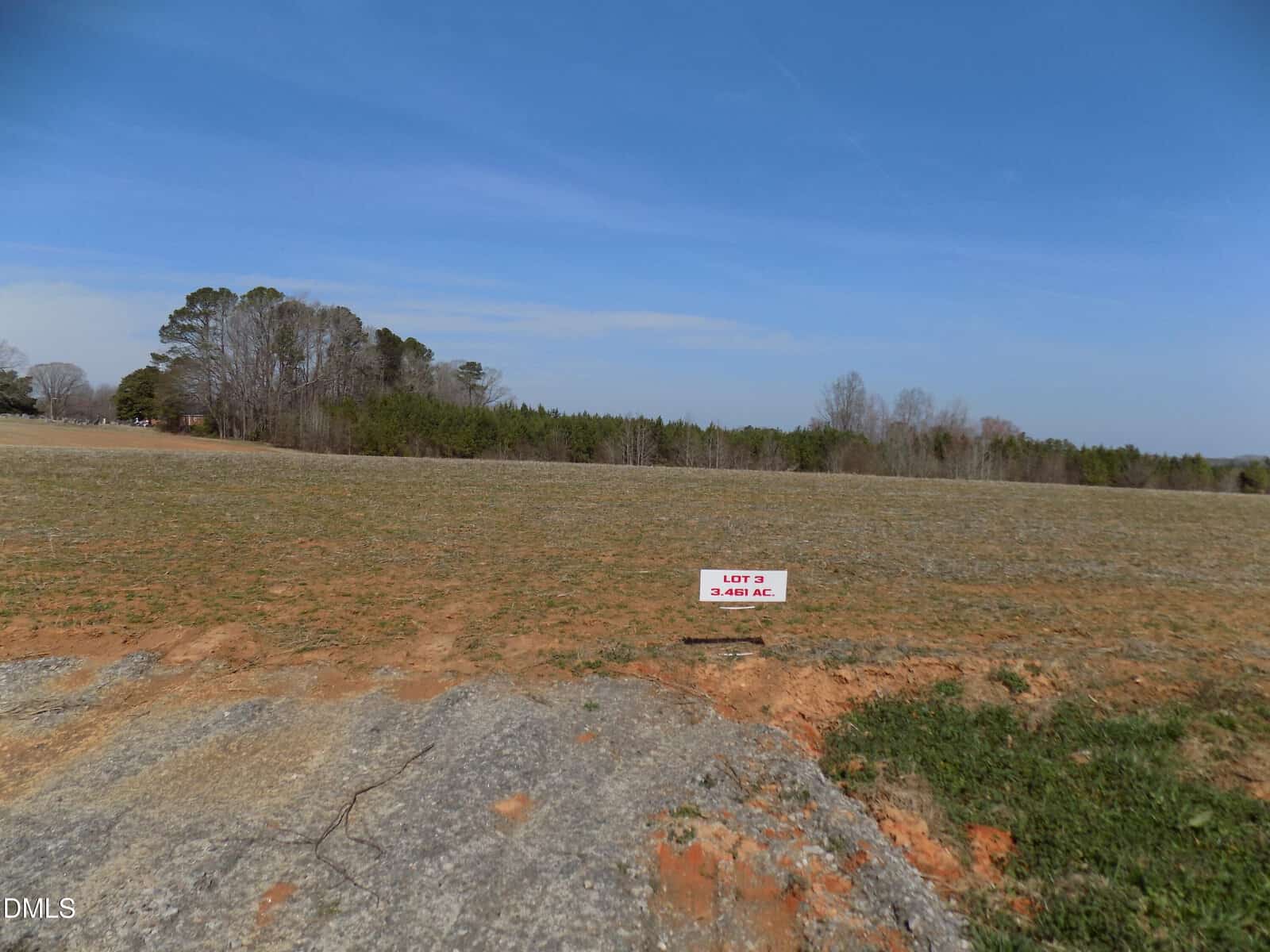A grassy, mostly flat open lot at 000 Sutton Road, Louisburg, with a small white sign reading “Lot 2, 2.45 AC” stands in the center; trees and blue sky are in the background.