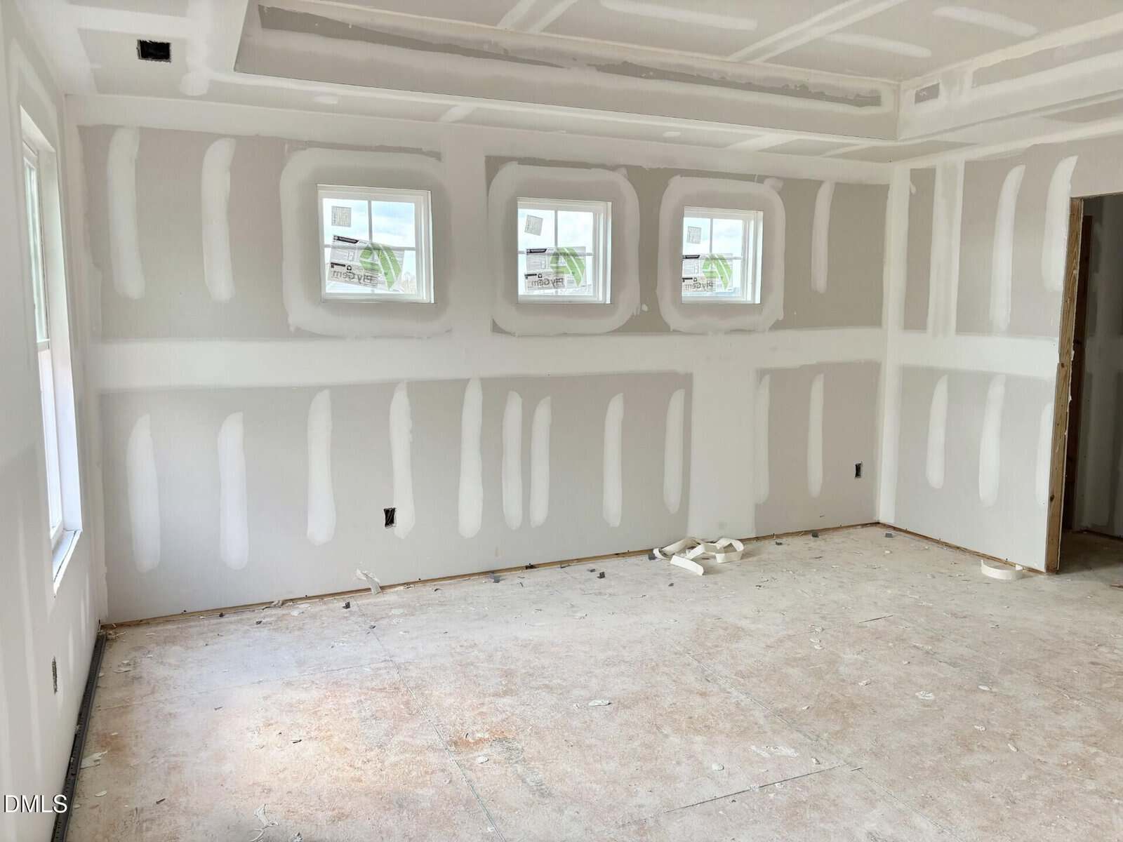 Unfinished room with drywall installed, showing white joint compound on seams and screw holes. Four square windows and one open doorway are visible. Floor is bare with some debris, and ceiling has visible drywall seams.