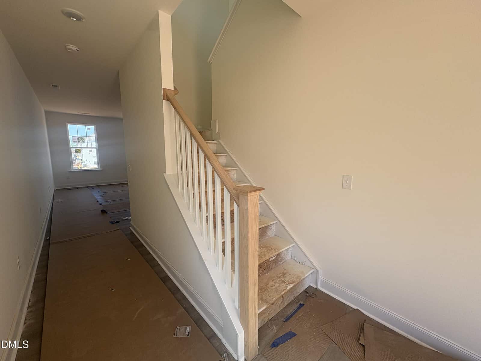 A staircase with a wooden handrail and white balusters leads up from a hallway in a house under construction. The floor is covered with protective cardboard, and walls are painted light beige. Natural light comes from a window at the far end.