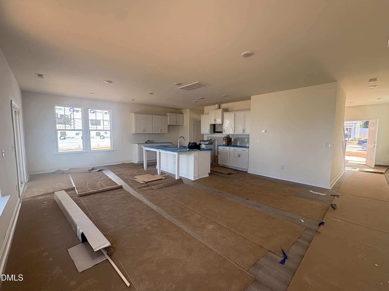 Open-concept kitchen and living area in a new home under construction, with protective paper covering the floors and uninstalled materials on the ground. White cabinets and a kitchen island are visible, with sunlight streaming through windows.