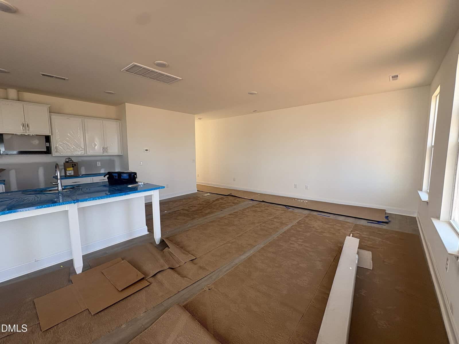Unfurnished room with white walls and covered floors, a kitchen area with white cabinets and a blue-covered countertop, and large windows letting in natural light. Construction materials and cardboard cover the floor.