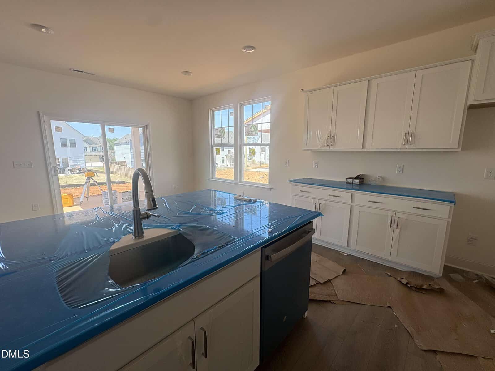 A modern kitchen with white cabinets, a blue-covered island with a sink, and large windows letting in sunlight. Some construction materials and cardboard are on the floor, indicating the space is newly built or renovated.