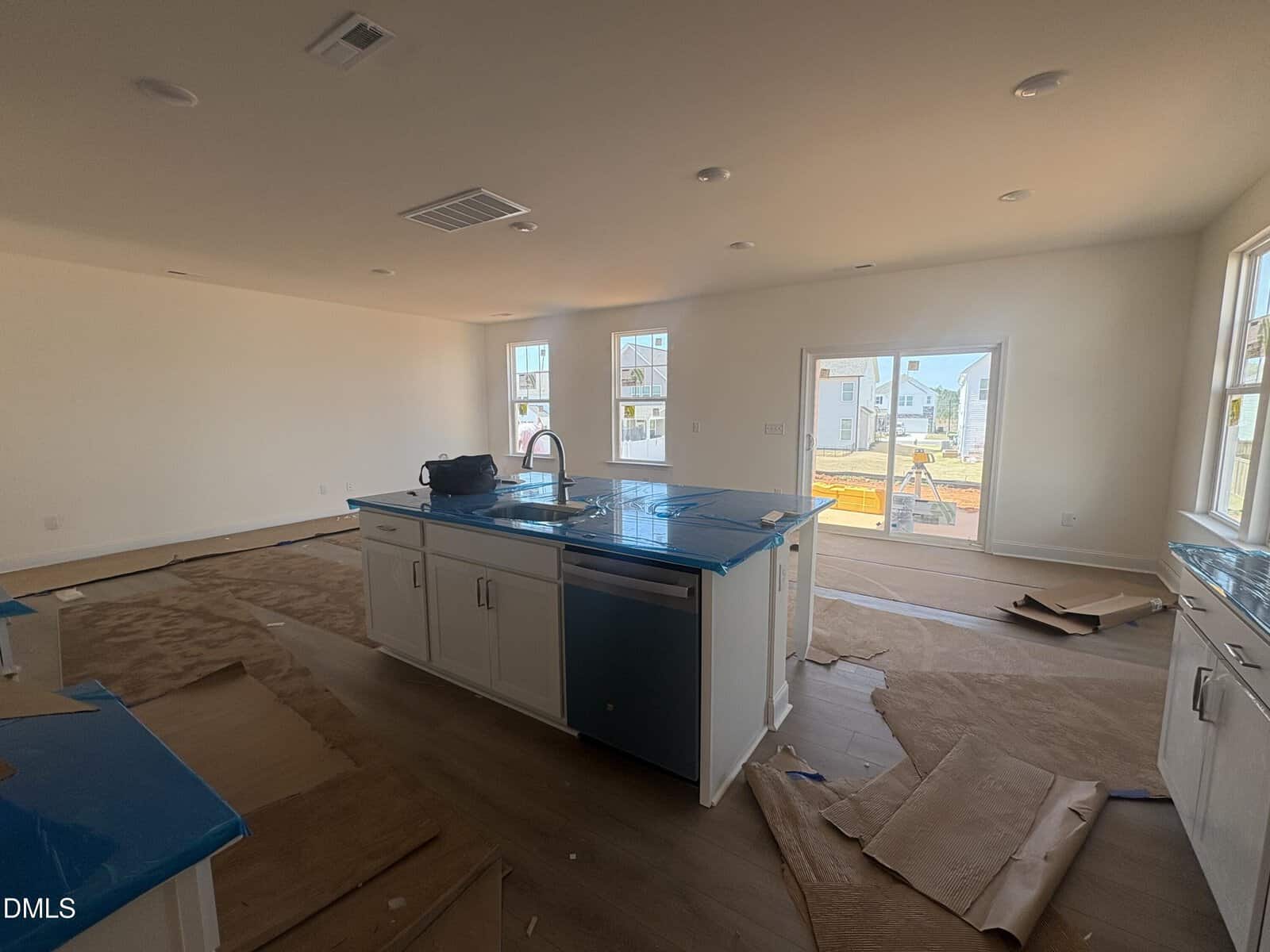 Unfinished kitchen with an island, white cabinets, protective blue film on counters, brown paper covering the floor, and bright windows letting in natural light. Construction materials are visible outside through the windows.