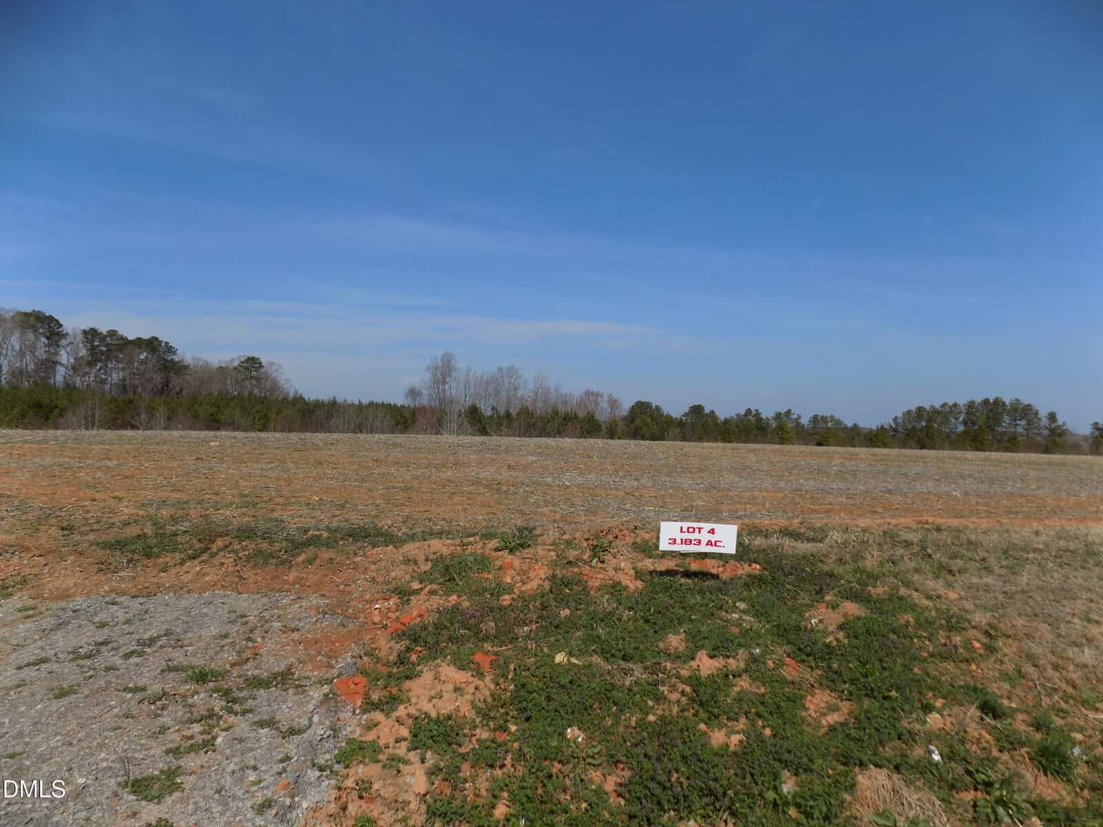 A mostly bare, open field with sparse grass and dirt patches under a blue sky stretches across 0000 Sutton Road, Louisburg. A small sign reads “Lot 4, 3.192 AC.” Trees line the horizon in the background.