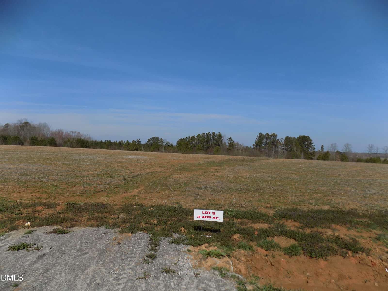 A grassy, open field along 00000 Sutton Road in Louisburg features a small sign reading “LOT 5, 2.4695 AC.” Trees line the background under a clear blue sky.
