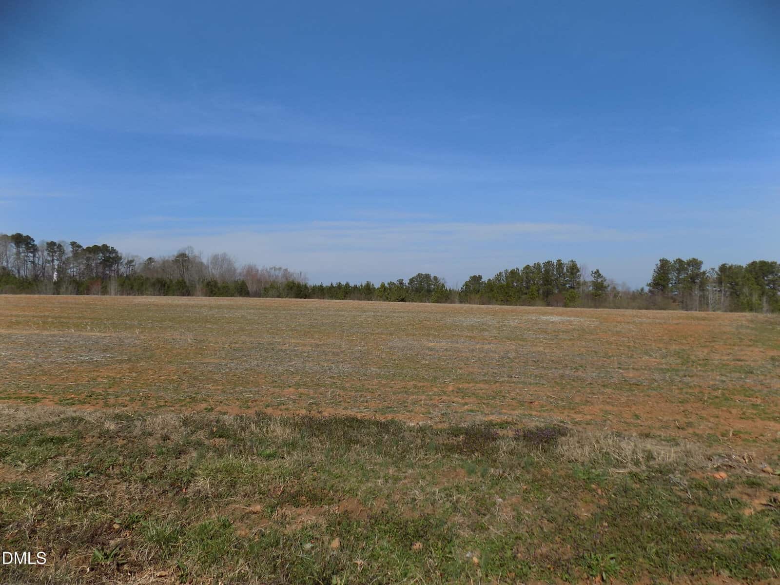 A large, open field with mostly bare, brownish soil and sparse grass. Trees line the horizon under a clear blue sky. No structures or people are visible.