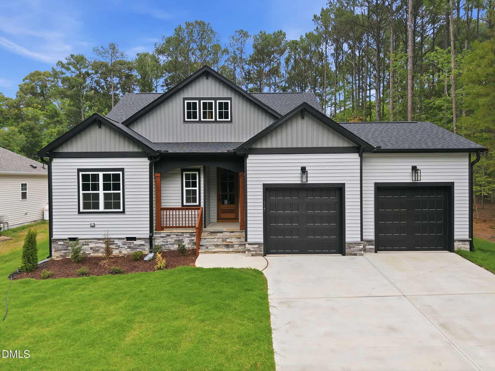 A modern single-story house at 117 Deer Cross Drive in Louisburg features gray siding, black trim, two black garage doors, a front porch with wooden railing, stone accents, a concrete driveway, green lawn, and trees in the background under a blue sky.