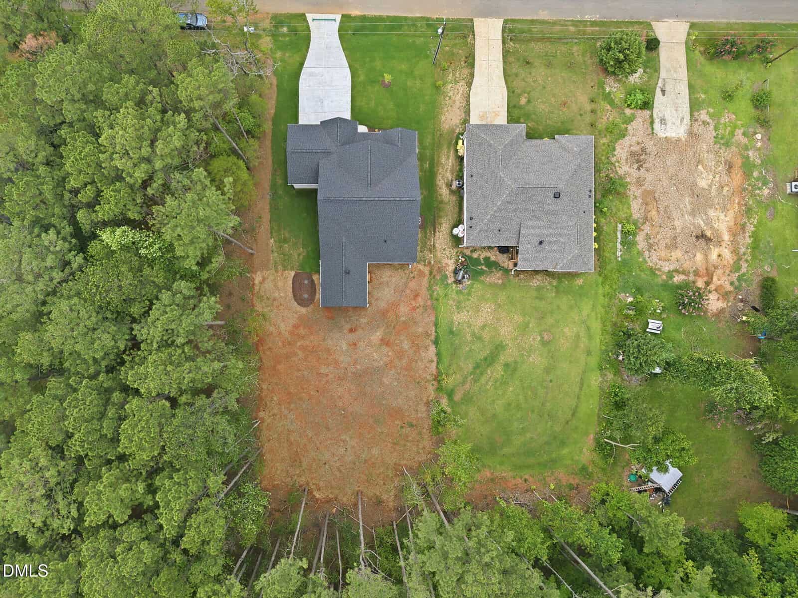 Aerial view of two houses with driveways, one lot with bare earth, grassy yards, and dense trees on the left and bottom edges.