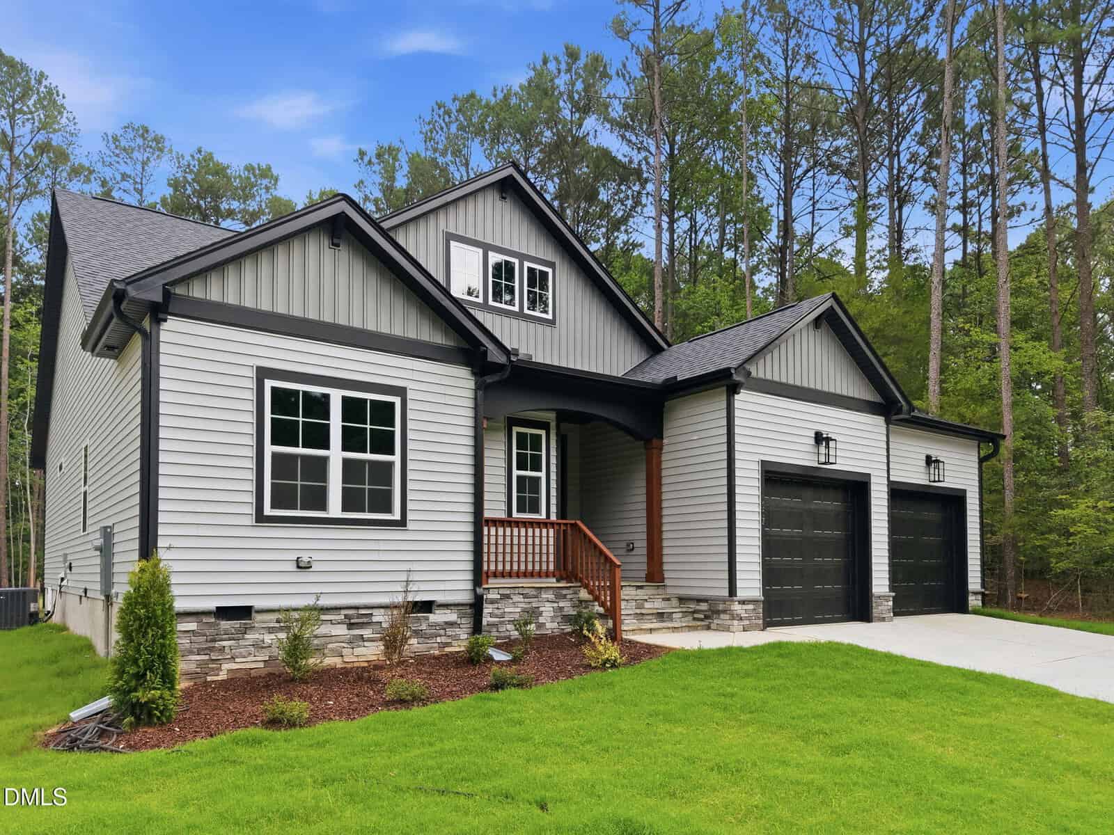 Modern gray house with black accents, double garage, covered porch with wooden steps, landscaped front yard, and surrounded by tall trees under a blue sky.