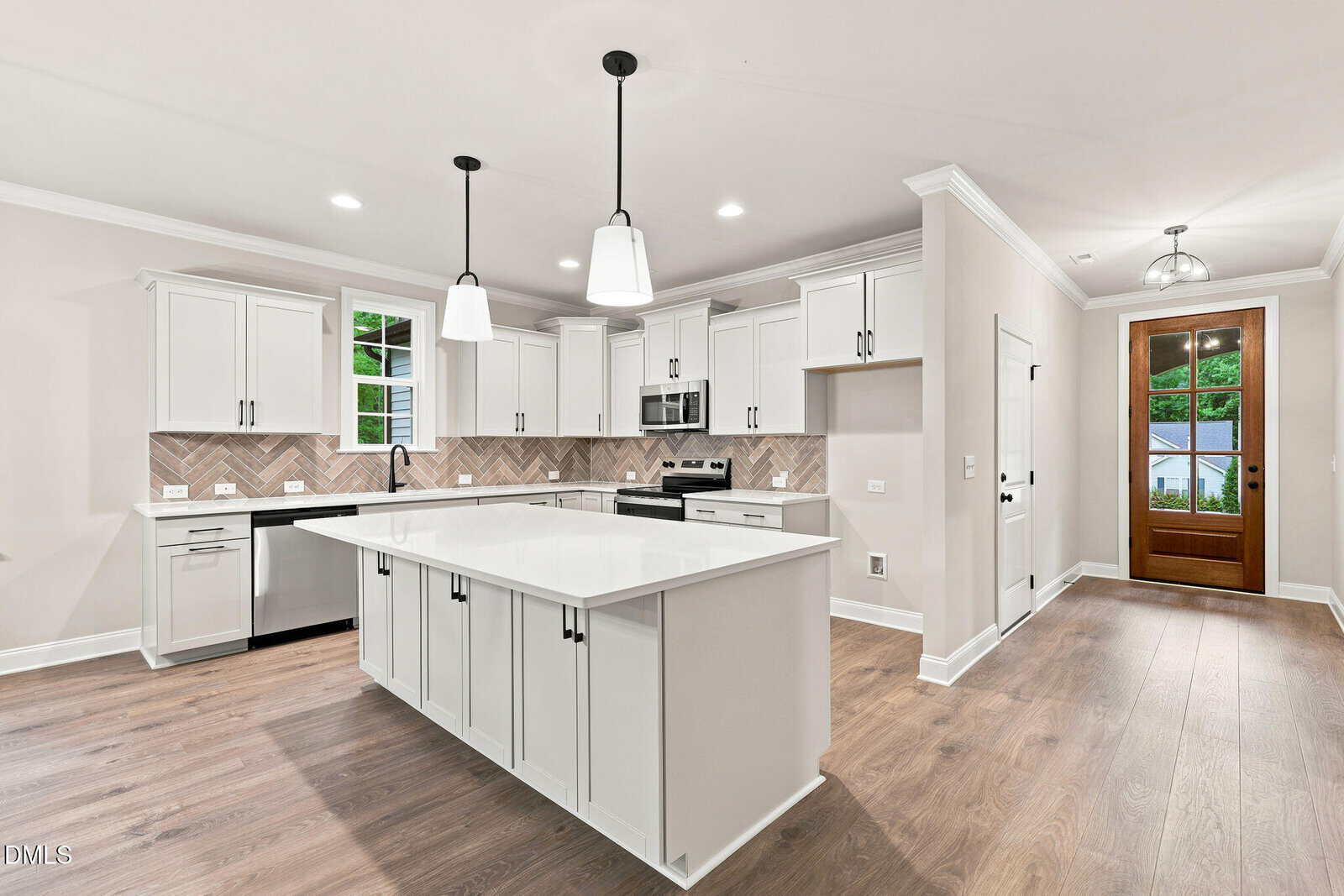Modern kitchen with white cabinets, island with white countertop, stainless steel appliances, herringbone backsplash, pendant lights, wood floors, and a glass door leading outside.