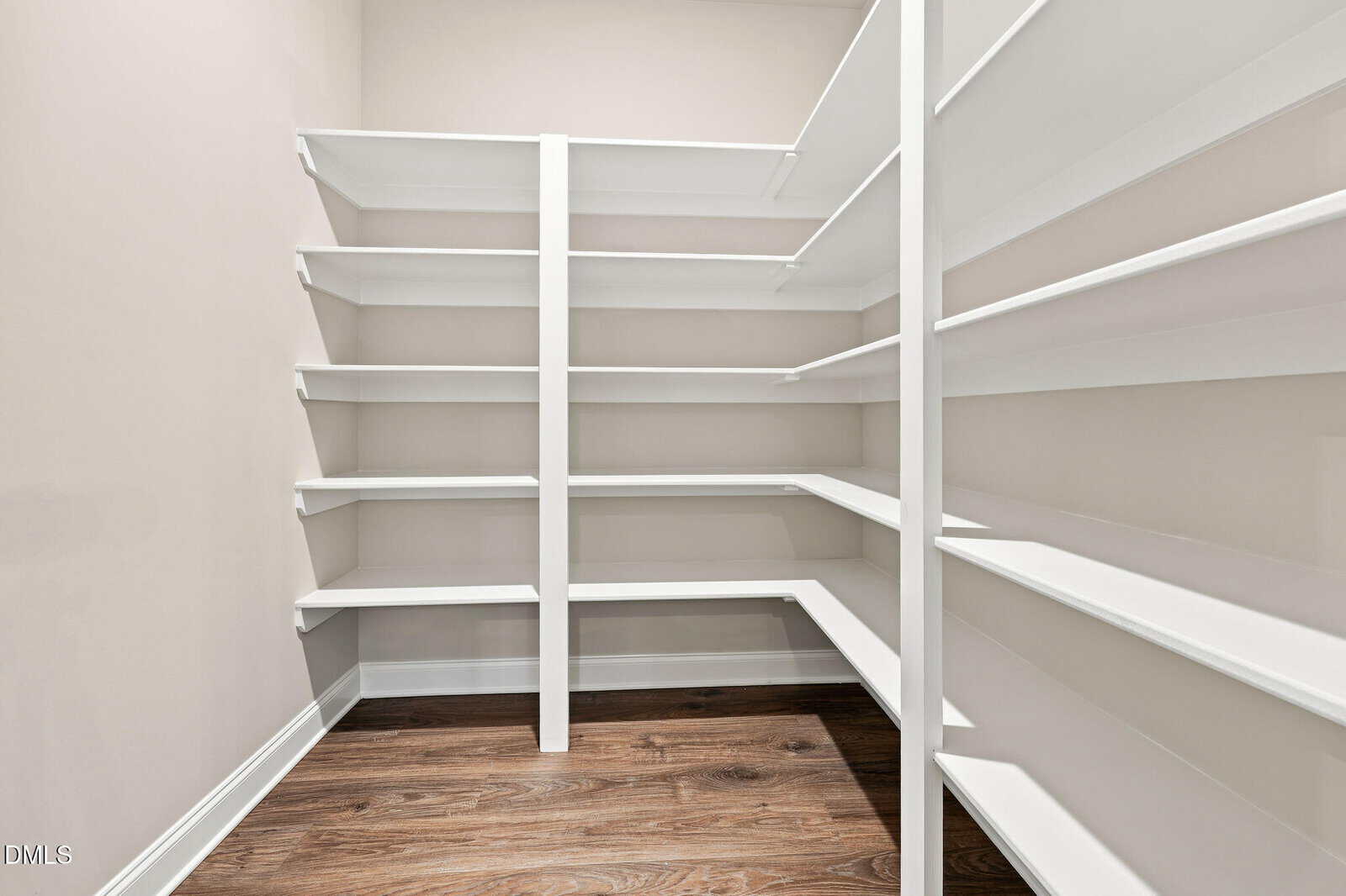 A walk-in pantry with white built-in shelves on three walls and a wood floor, all empty and brightly lit.