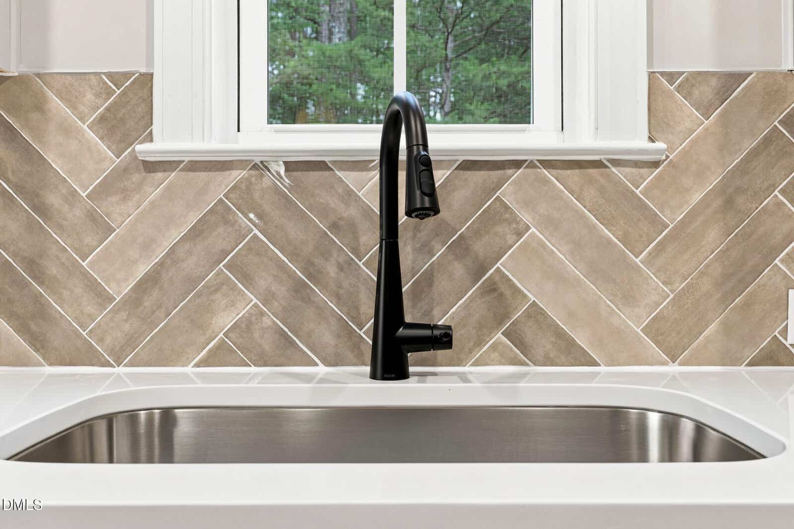 A modern black kitchen faucet over a stainless steel sink, set against a beige herringbone tile backsplash with a window above.