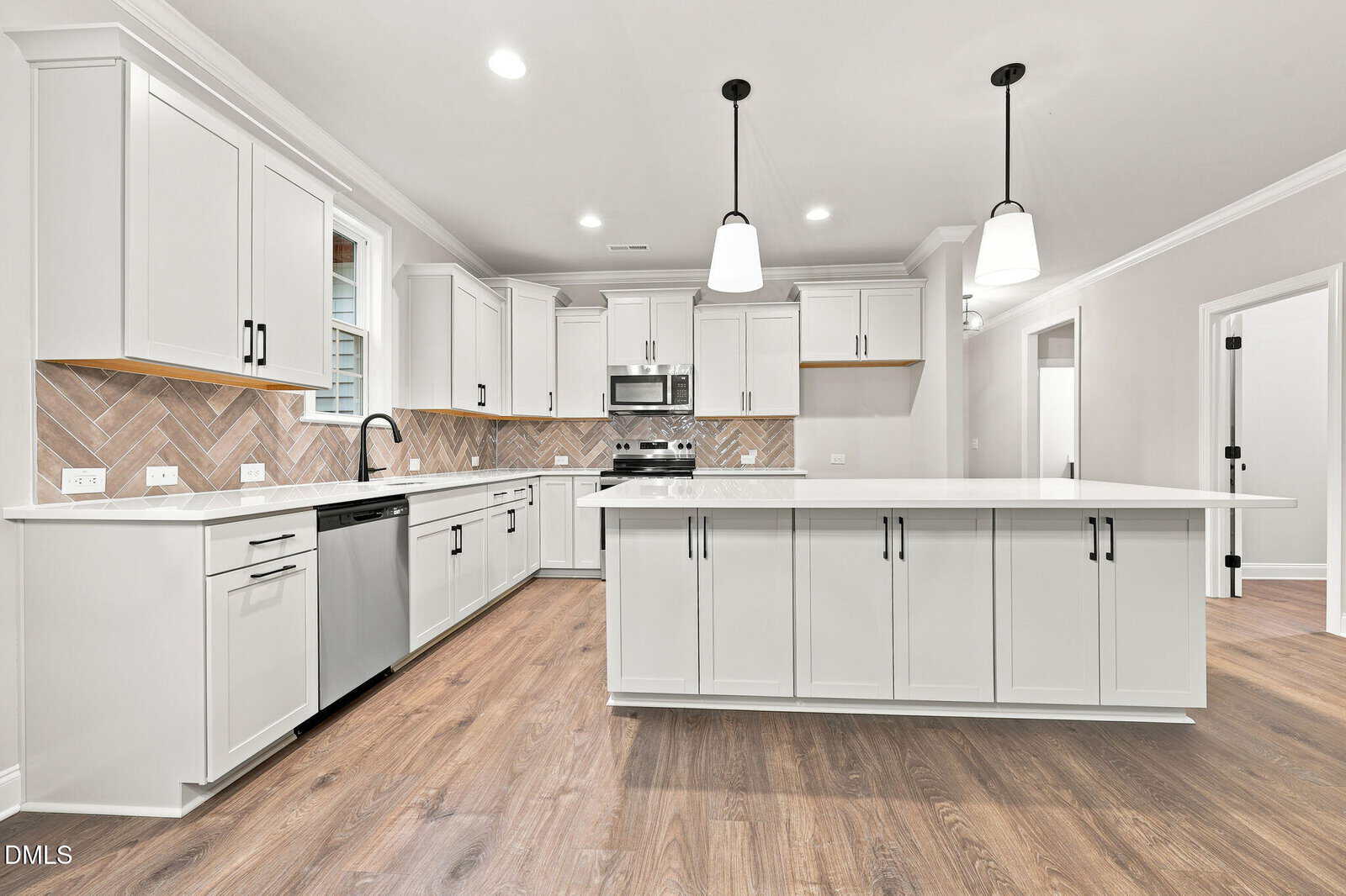 Modern white kitchen with large island, stainless steel appliances, herringbone tile backsplash, pendant lights, and wood-look flooring. Cabinets have black handles and the counters are bright and spacious.