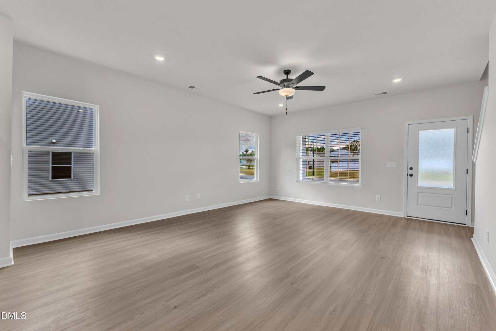 Empty, spacious room with light-colored wood flooring, white walls, three windows, a ceiling fan with a light, recessed ceiling lights, and a white door with frosted glass. The room appears well-lit and modern.