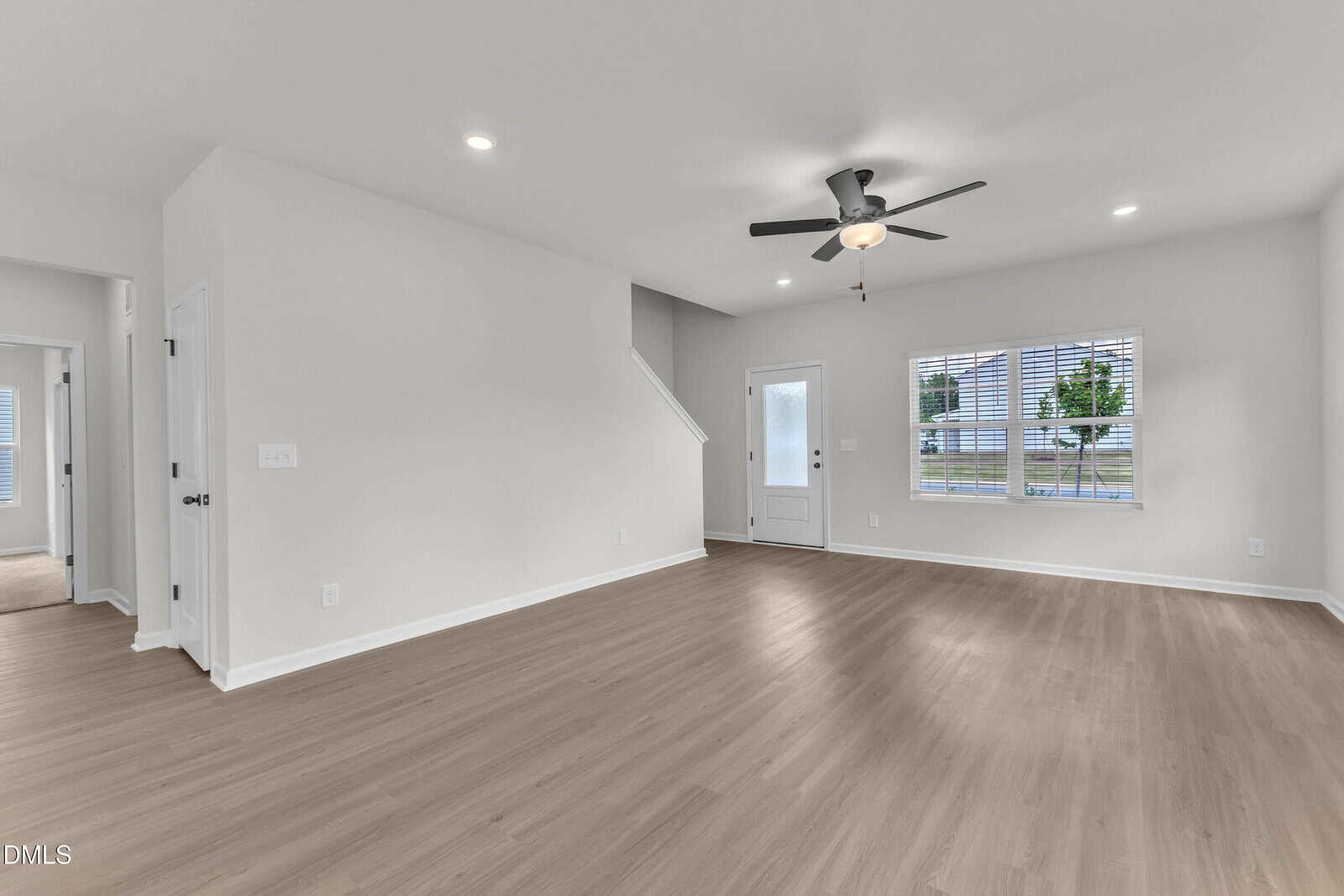 A bright, empty living room with light wood flooring, white walls, recessed lighting, a ceiling fan with light, a window, and a door leading outside. Stairs and a hallway are visible on the left side.