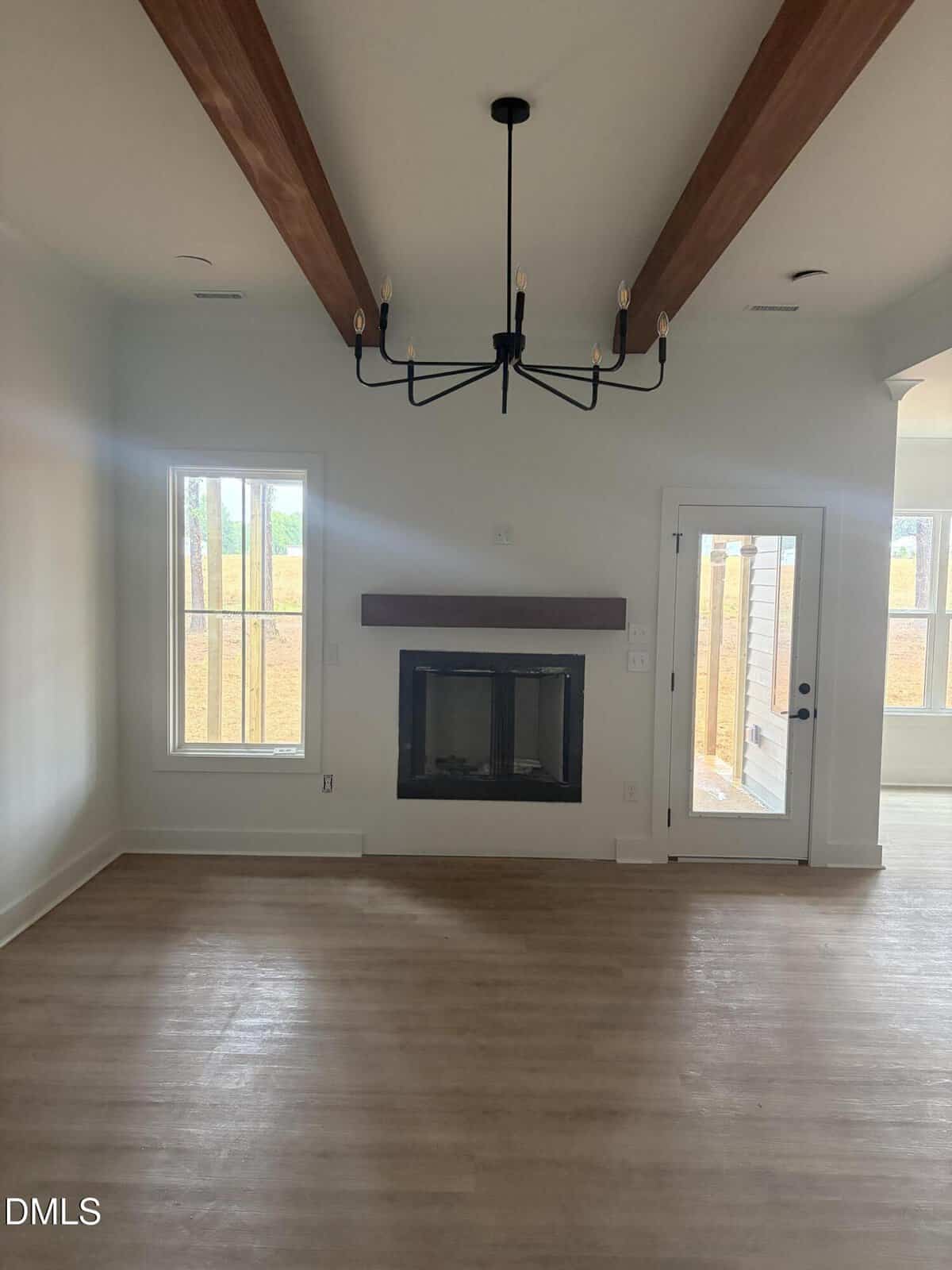 A modern, unfurnished living room with light wood floors, white walls, exposed wooden ceiling beams, a black chandelier, a fireplace with a wooden mantel, and windows and a glass door letting in natural light.