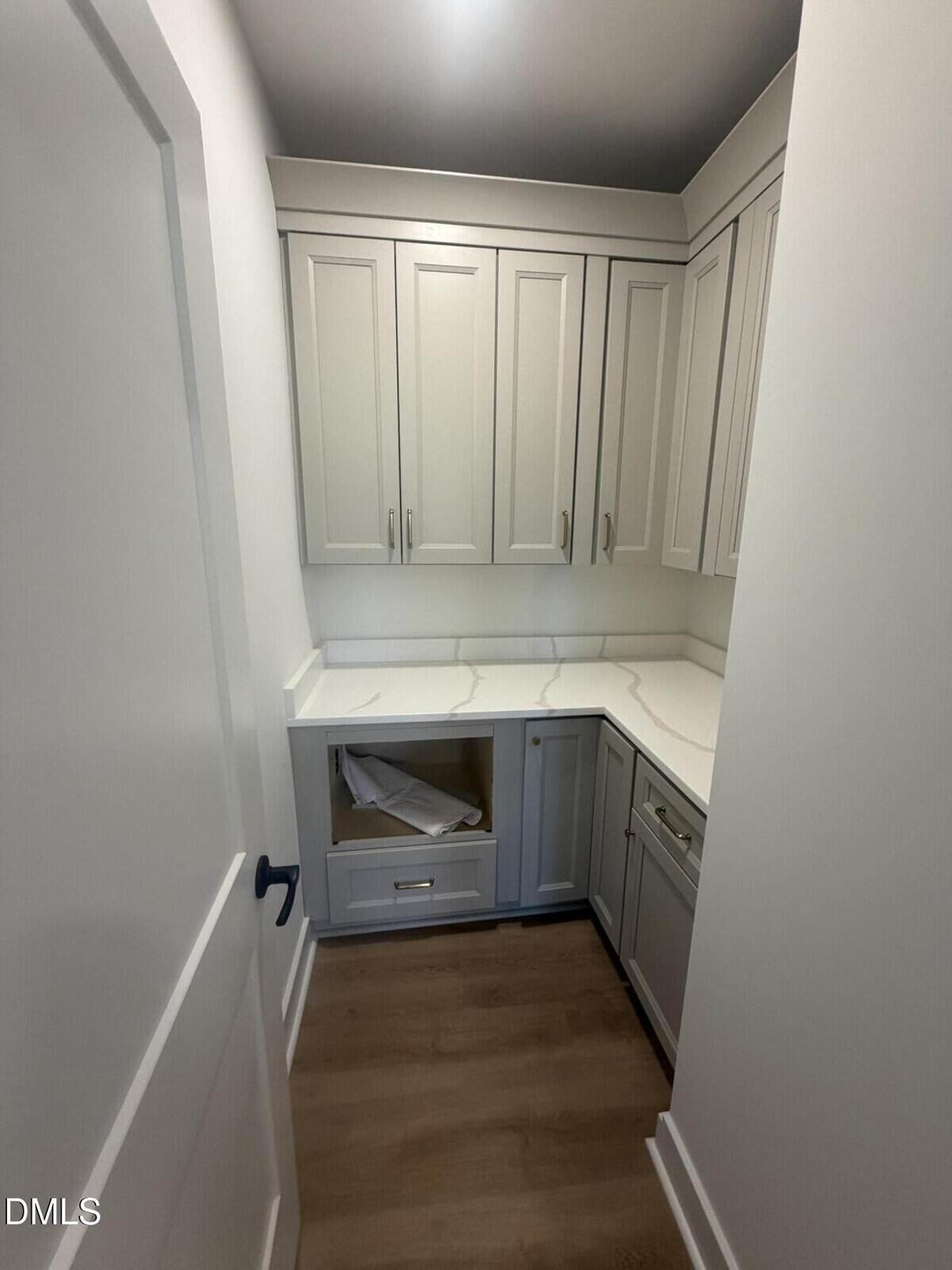 A small room with white cabinets, light wood flooring, white countertops, and an empty space for an appliance. The walls and door are also white.