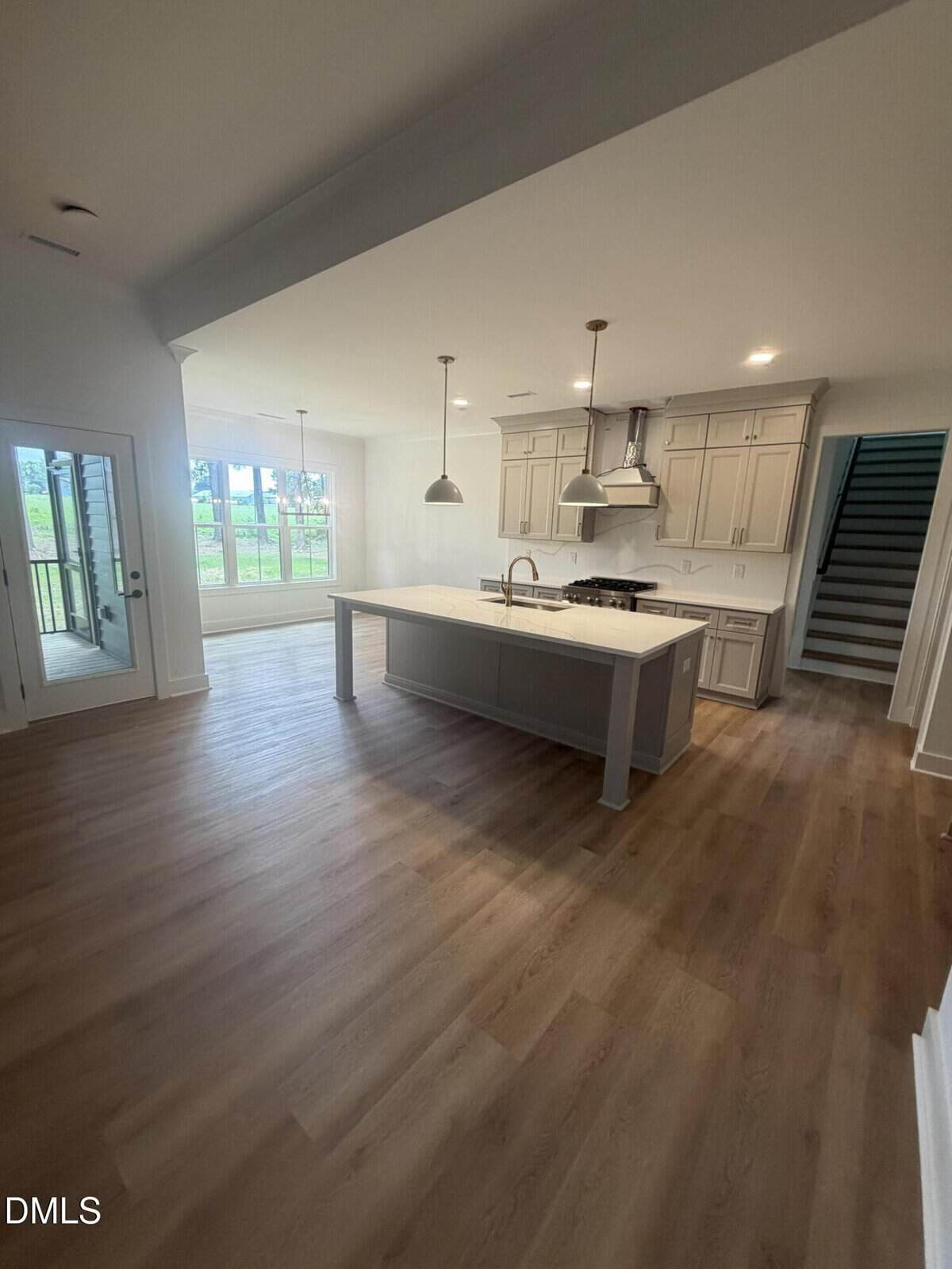 Modern kitchen with light wood floors, large island with sink, pendant lights, beige cabinets, and stainless steel appliances. Dining area by glass doors and stairs visible on the right. Bright and spacious layout.
