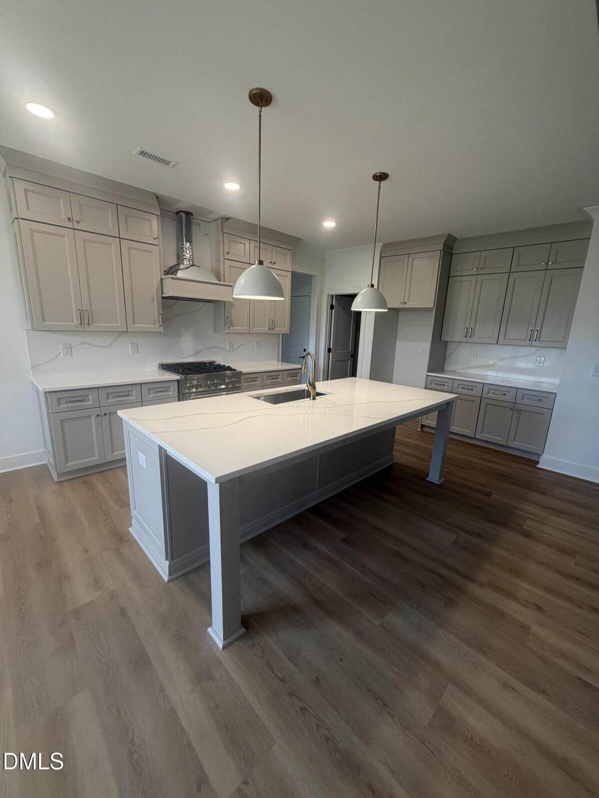 Modern kitchen with light gray cabinets, white countertops, a large island with a sink and gold faucet, two pendant lights, and wood-look flooring. Stainless steel range and hood are visible against the wall.